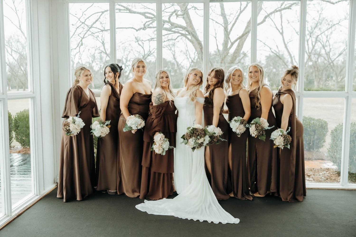 Wedding party poses in a panoramic shot with bridesmaids in brown dresses and white floral bouquets.