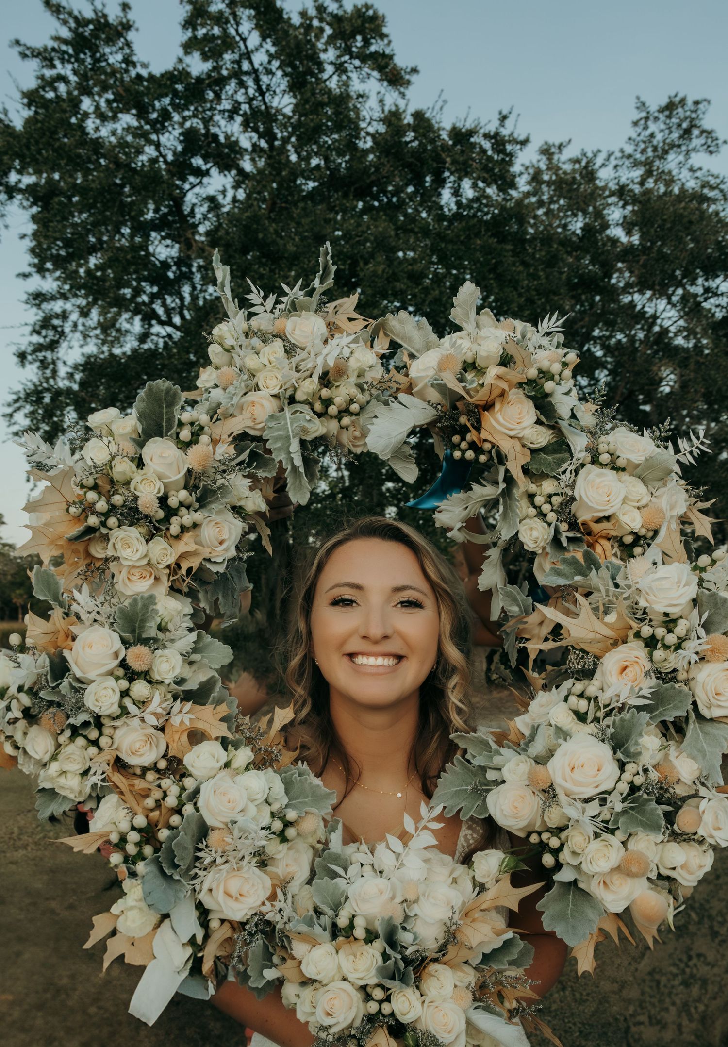 A beautiful floral wreath made of white roses and delicate blooms creates a circular frame against a natural outdoor setting.