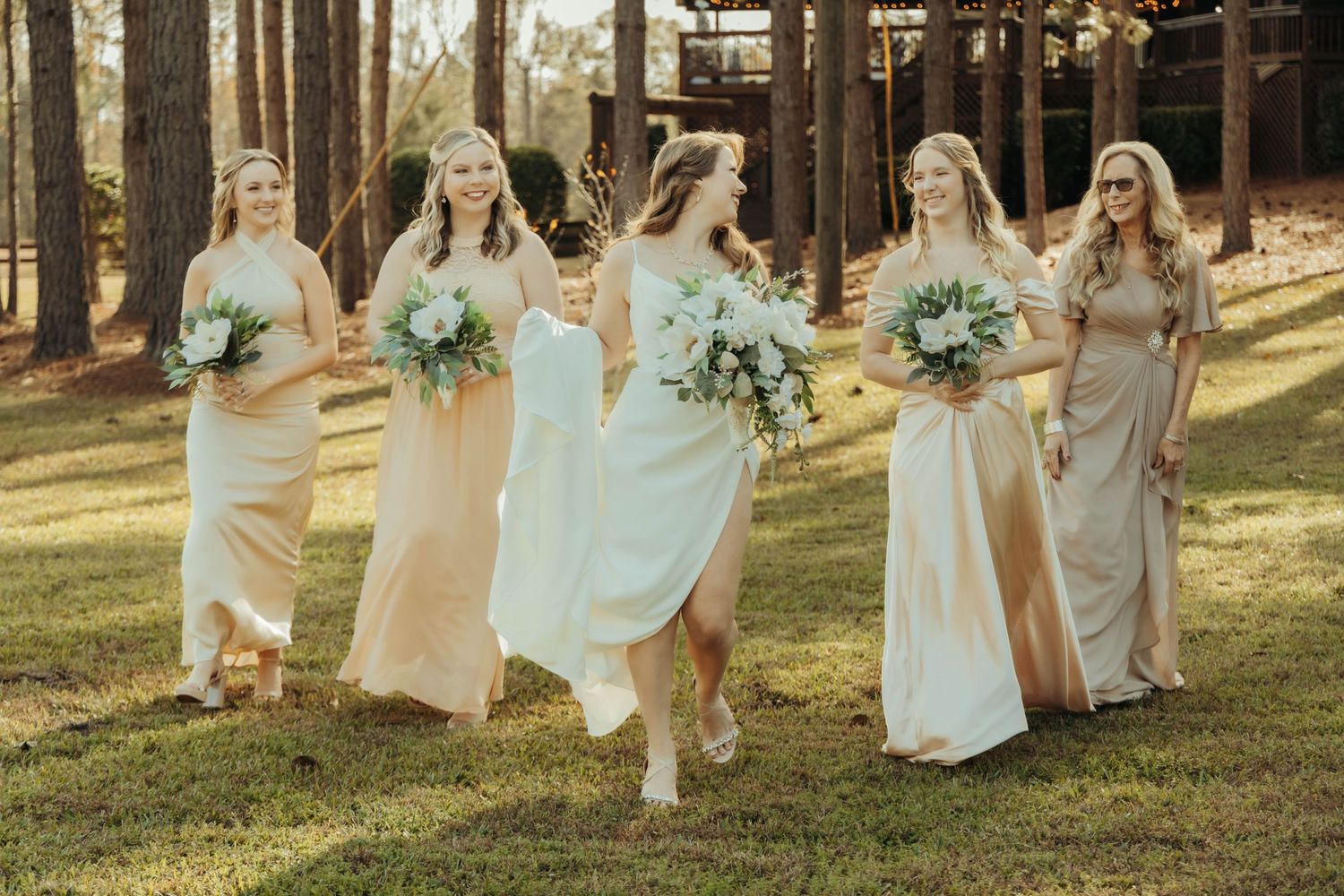 Bridesmaids in neutral dresses walking through a sunlit pine forest.