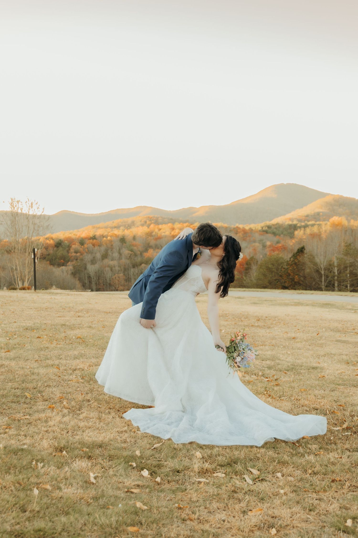A couple shares a romantic moment at sunset in a mountainous field with golden light and fall foliage in the background.
