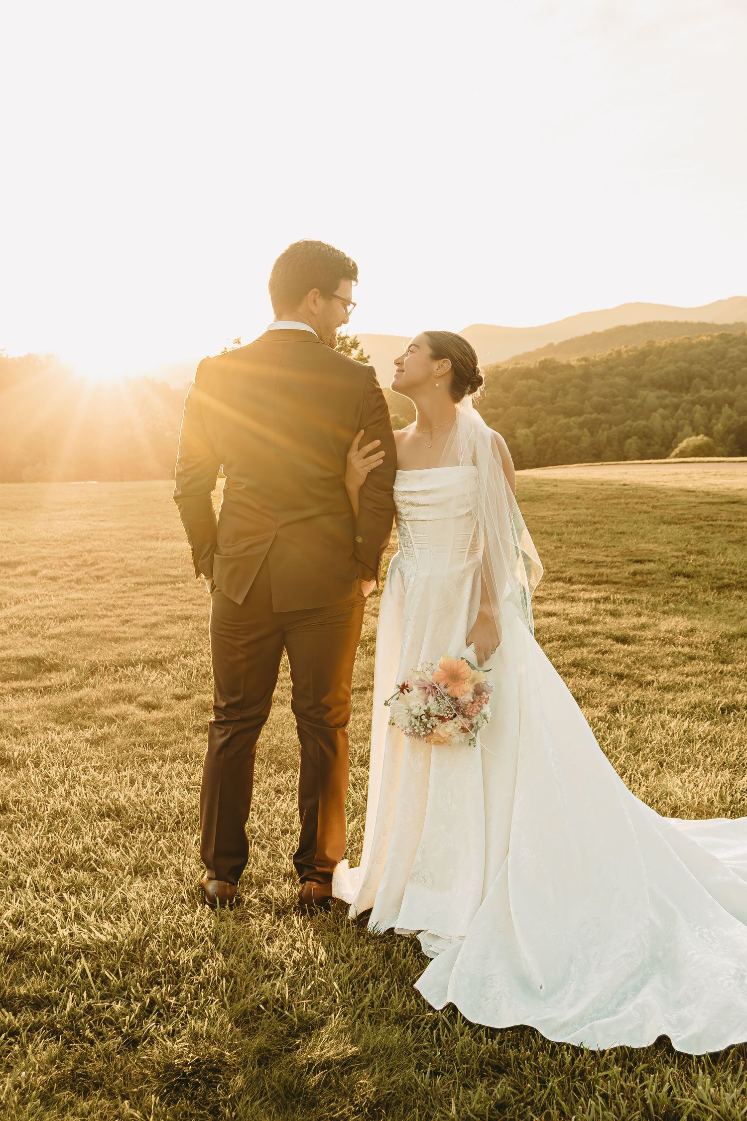 Wedding couple walking hand in hand through a sun-drenched meadow at sunset.