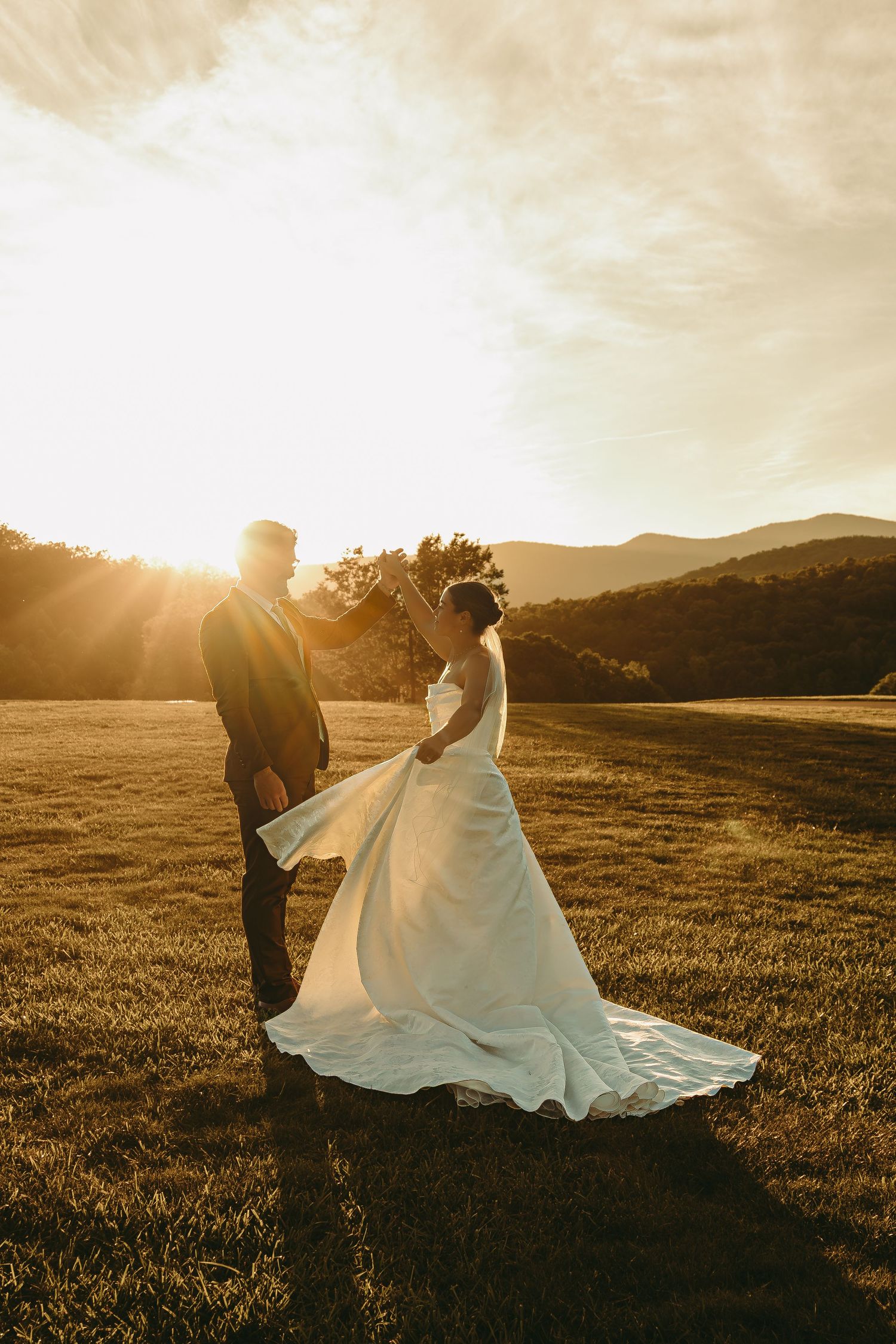 A wedding couple stands in silhouette against a golden sunset in a mountain meadow.