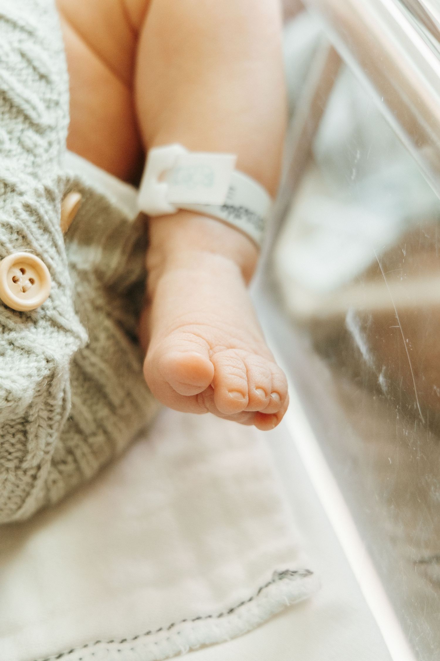 A tiny baby foot with a hospital ID bracelet peeks out from a knitted sweater against white bedding.