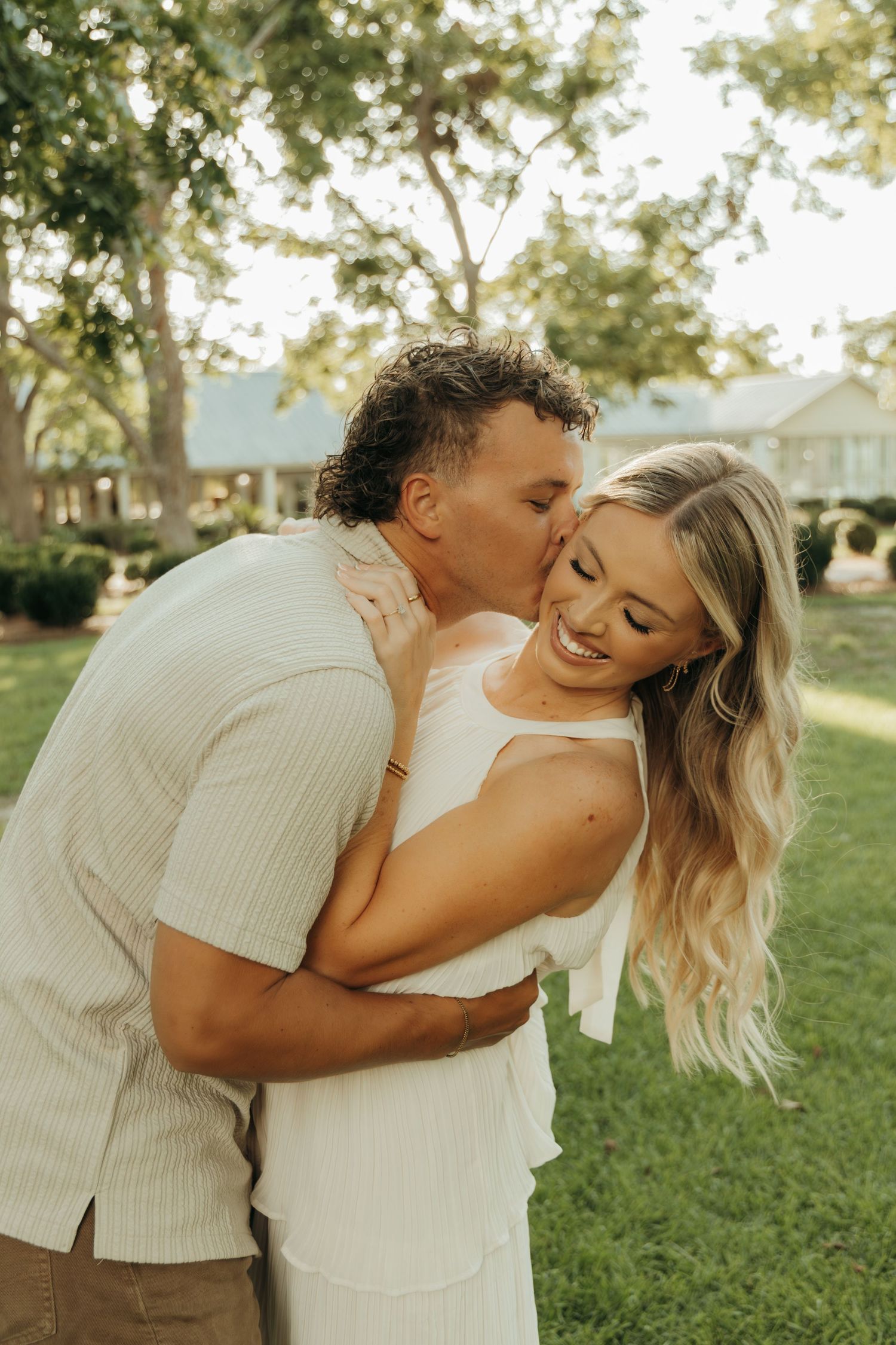 A romantic couple shares a tender moment outdoors, embracing and laughing in a sun-lit garden setting.