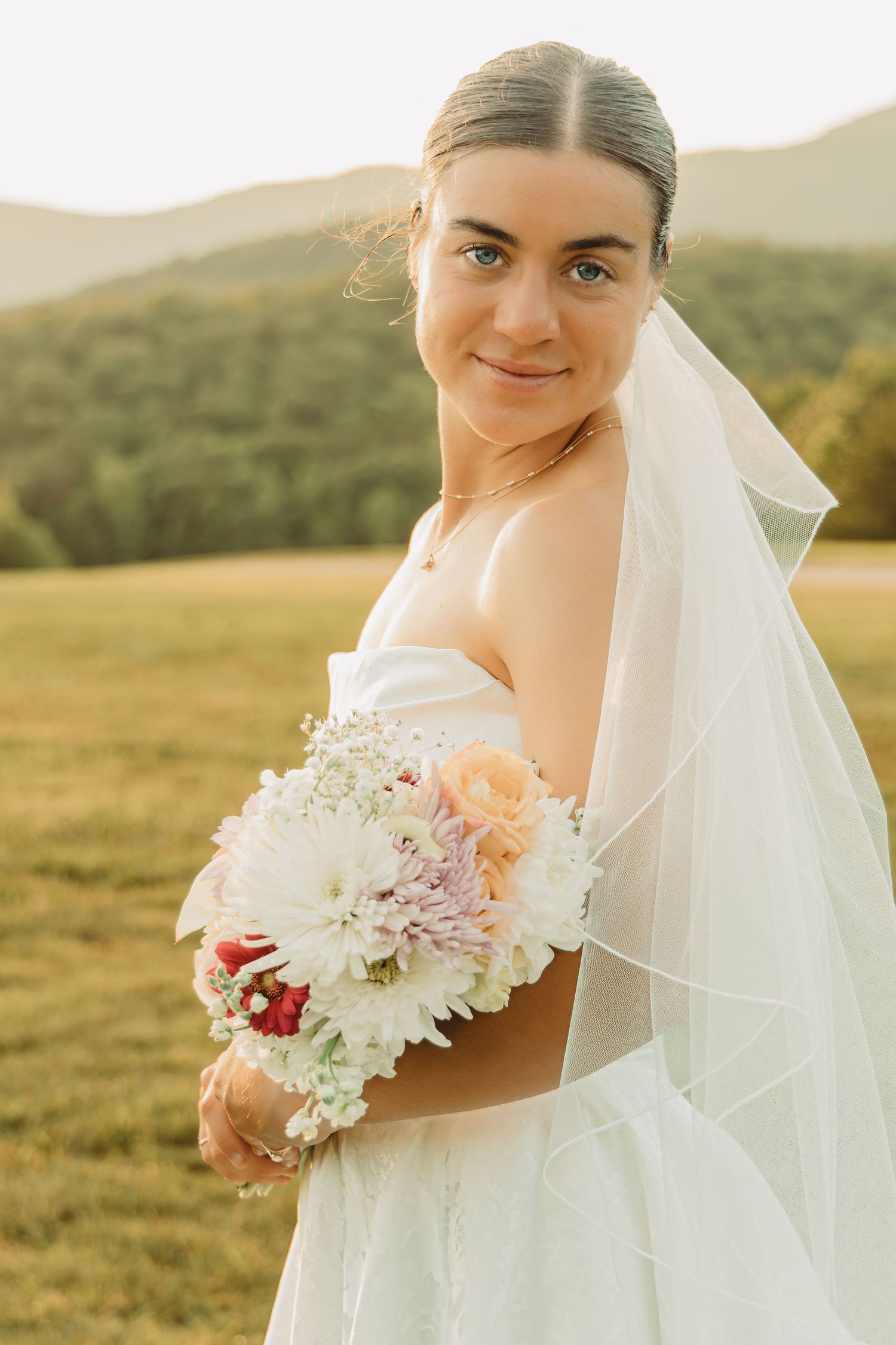 A bride in a white dress holds a colorful bouquet while posing in a sunlit meadow.