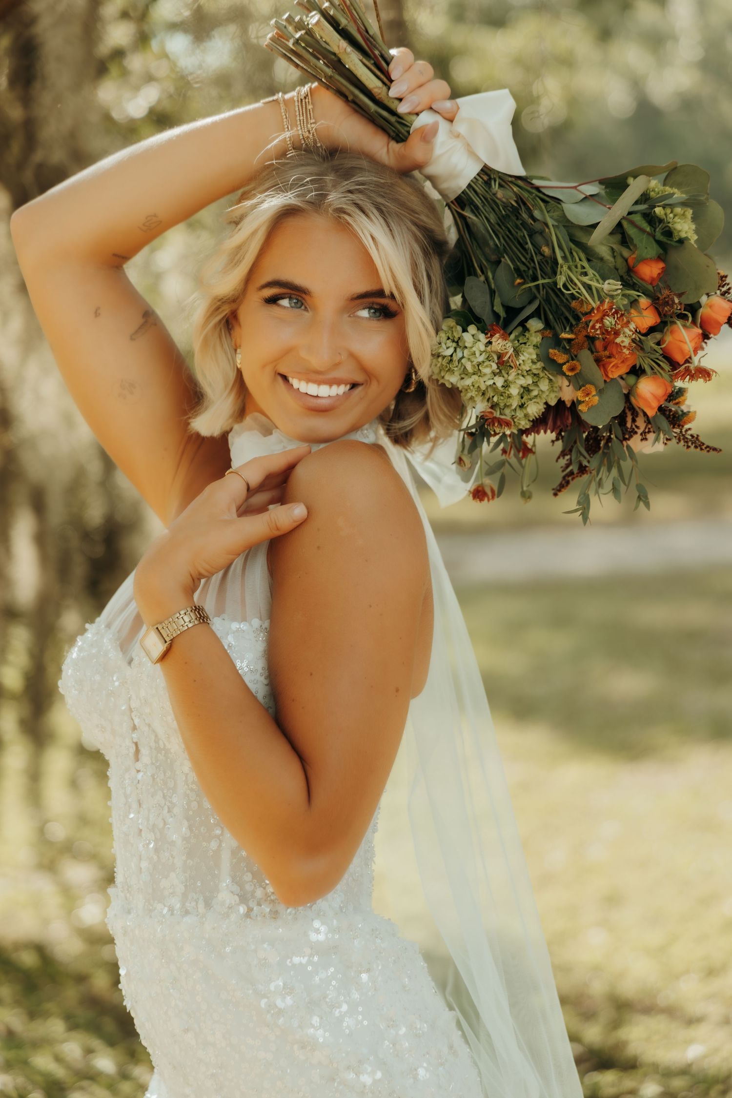 A person in a white sparkly wedding dress poses with a vibrant orange and green floral bouquet in a sunlit outdoor setting.
