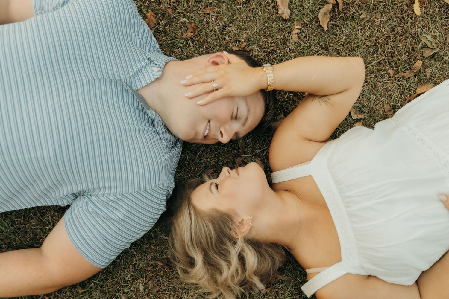 A romantic couple lying on grass together in a tender embrace wearing blue and white casual clothing.