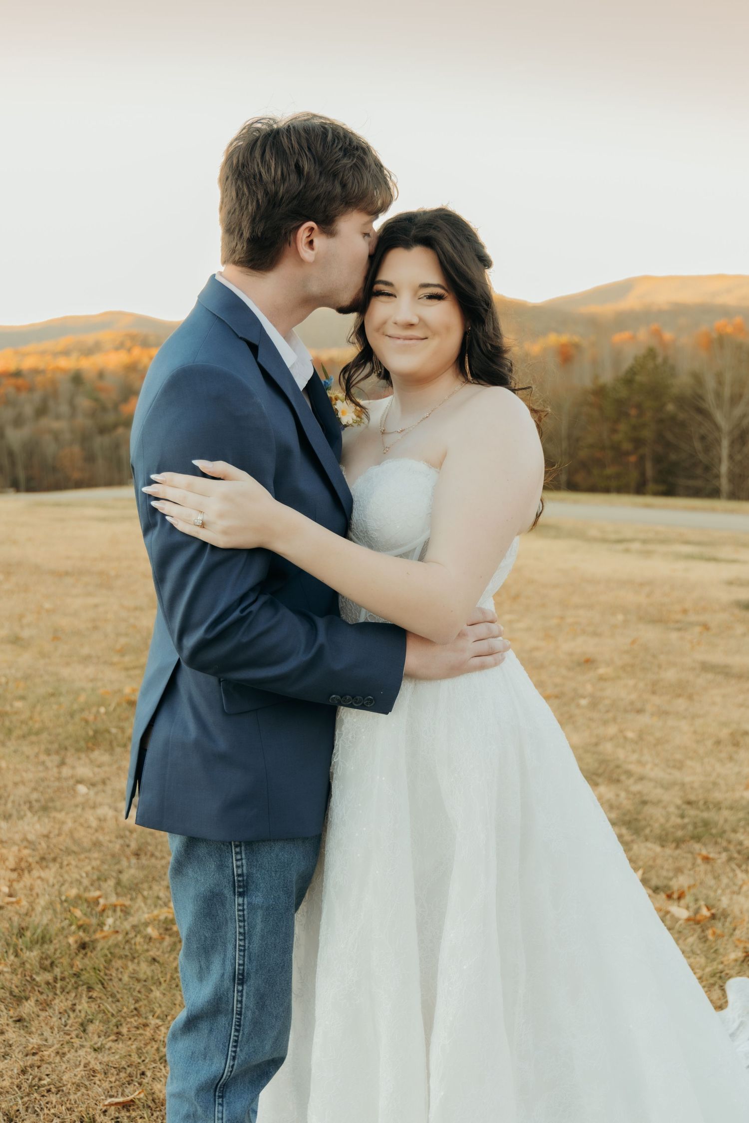 A couple embraces in a romantic autumn field with golden hills and trees in the background at sunset.