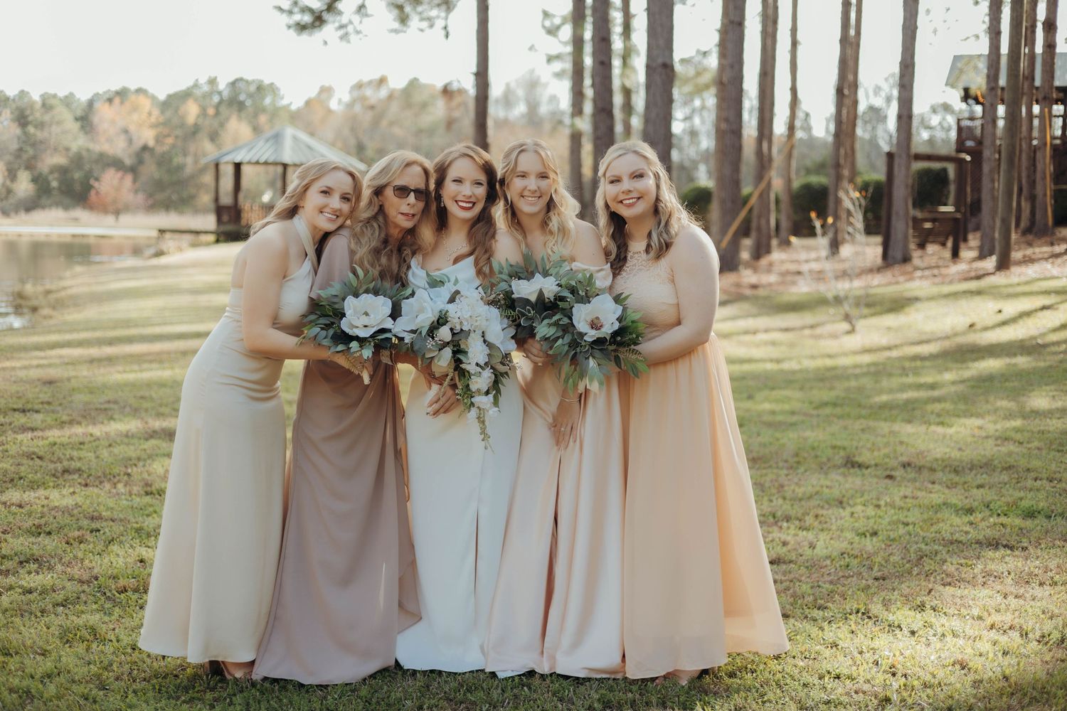 Wedding party in soft neutral colors gathered outdoors among tall pine trees.