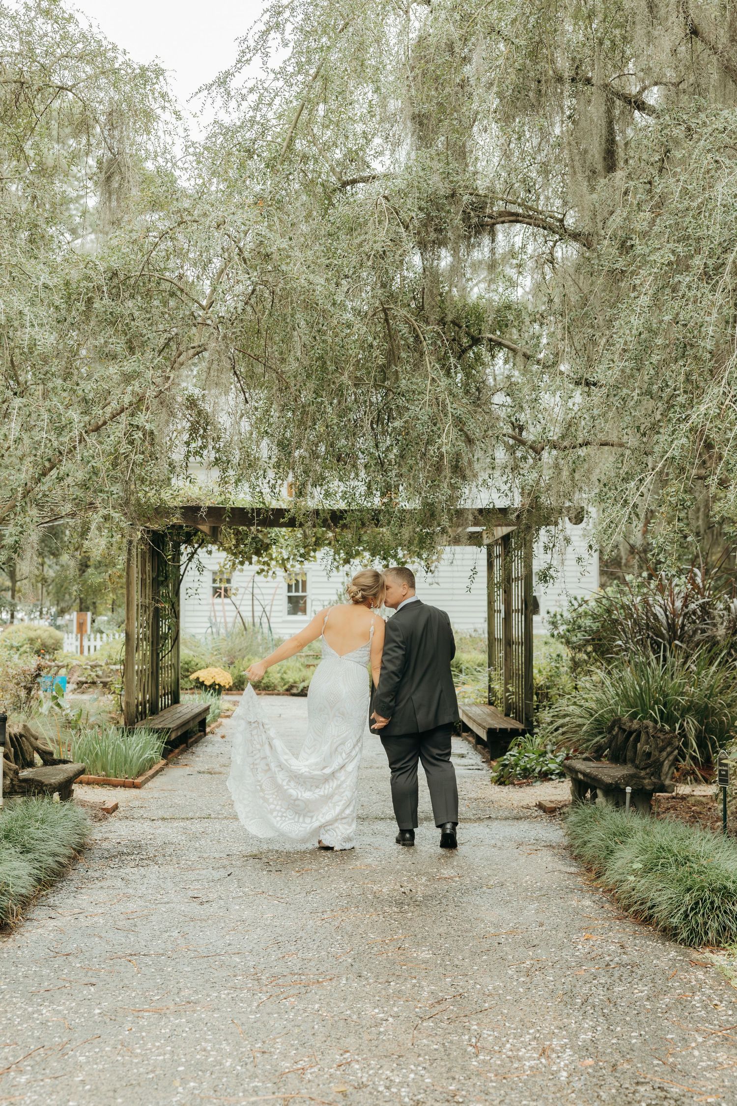 A romantic wedding couple shares intimate moments under weeping willow trees along a garden pathway.