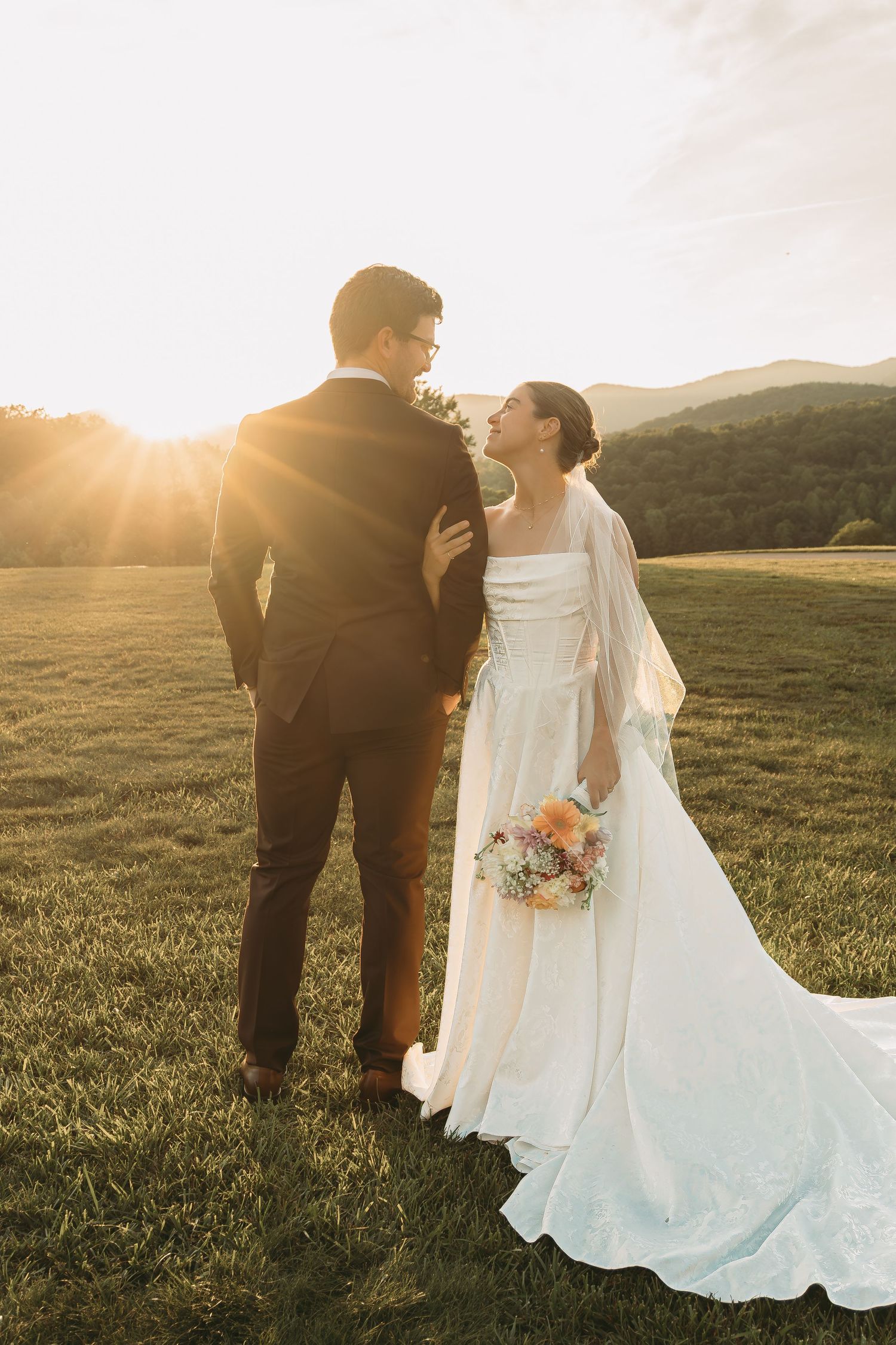 A couple in wedding attire shares an intimate sunset moment in a scenic mountain field.