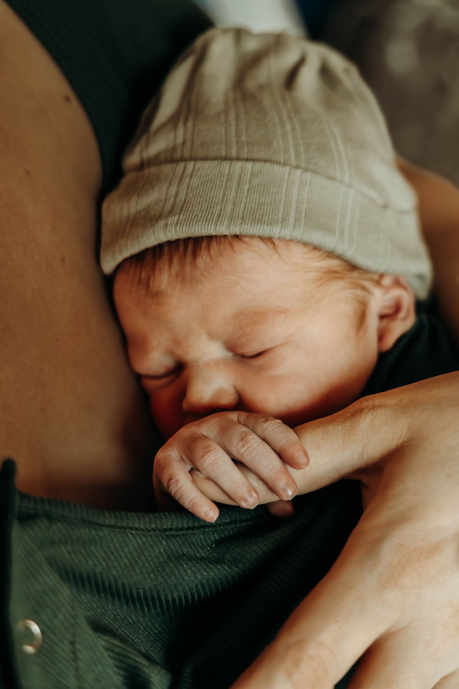 A sleeping newborn baby wearing a beige knit cap grasps an adult's hand while resting against their chest.