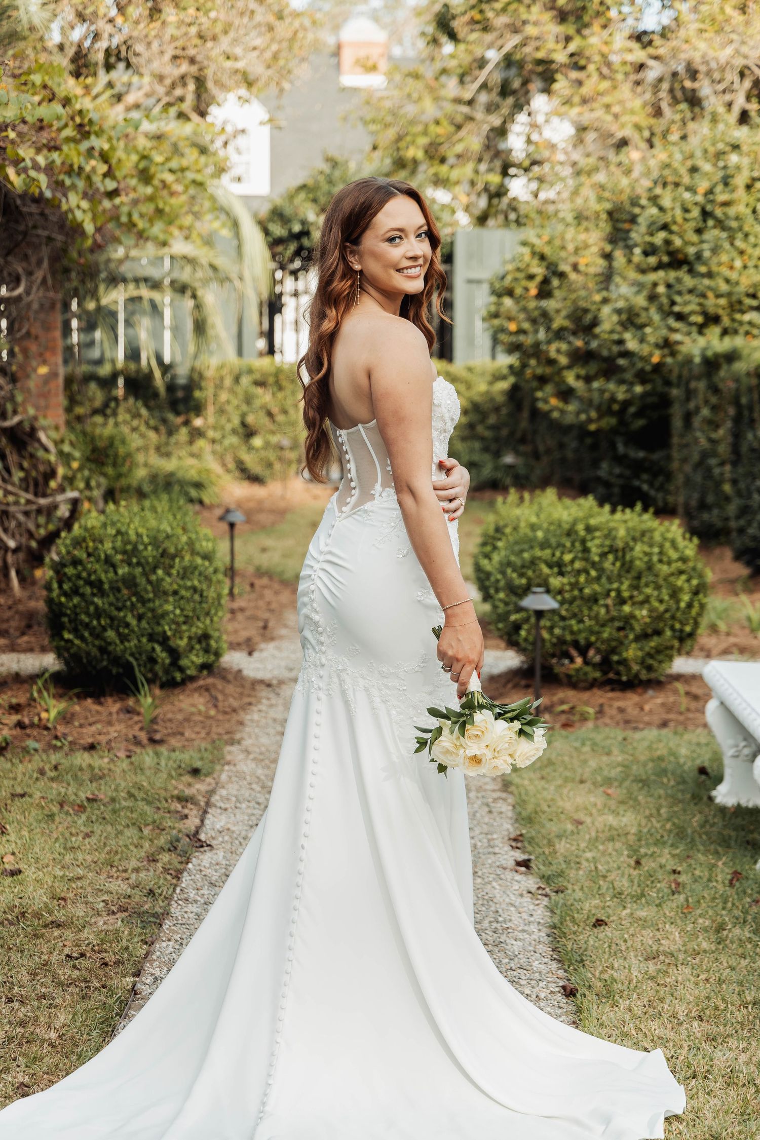 Elegant mermaid wedding dress with lace corset and flowing train shown in outdoor garden setting.