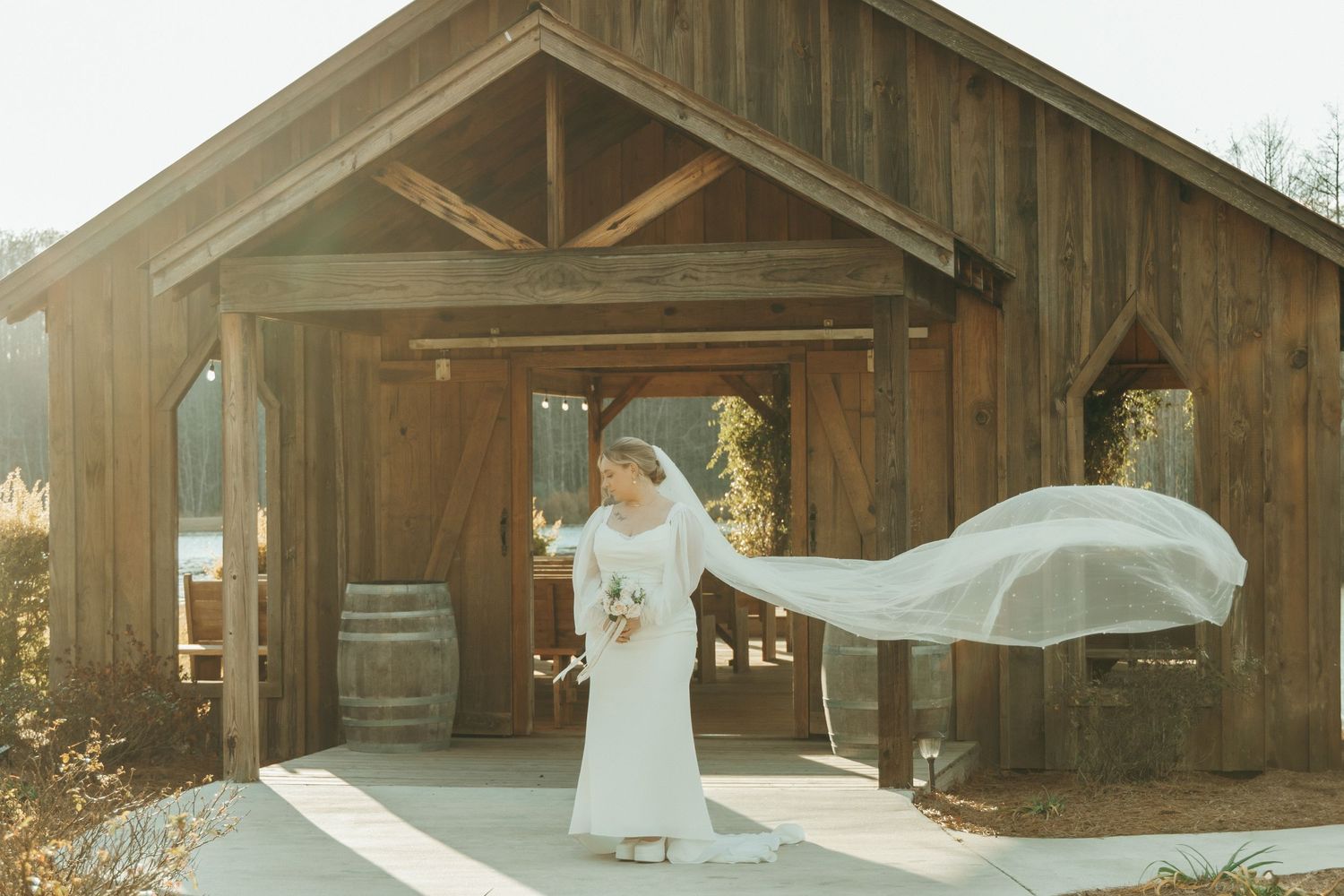 Flowing wedding veil catches wind in front of rustic wooden barn venue.
