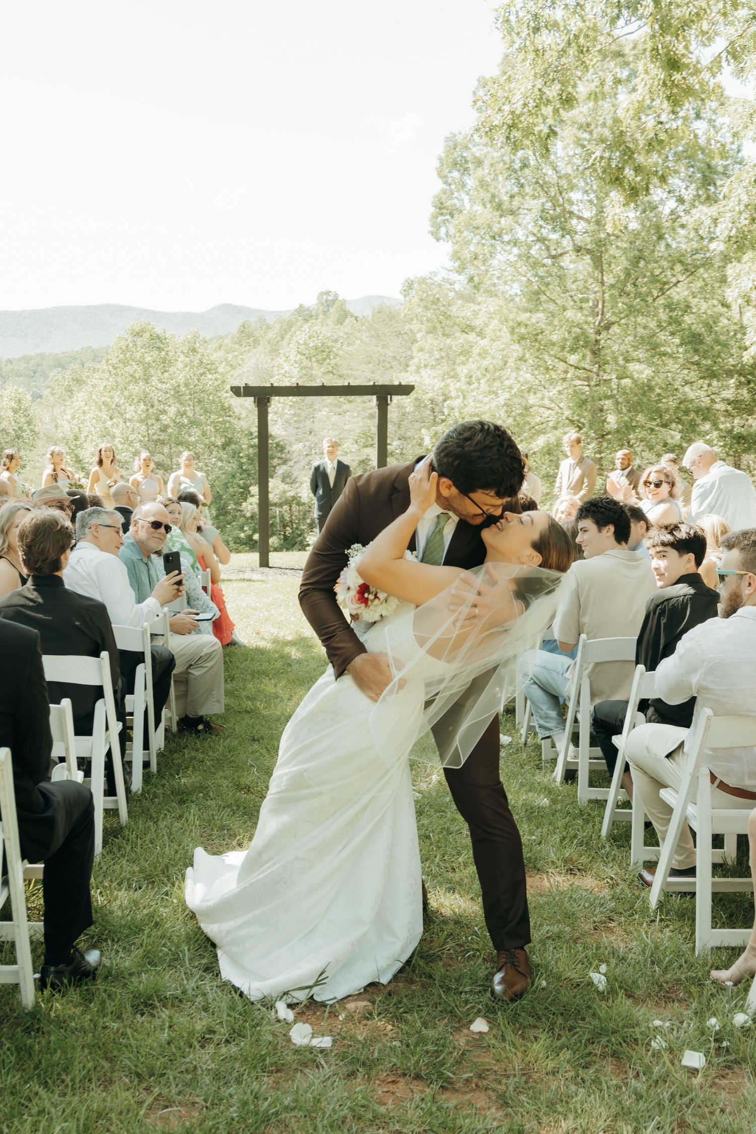 A romantic dip kiss between newlyweds at an outdoor mountain ceremony with guests seated on white chairs.