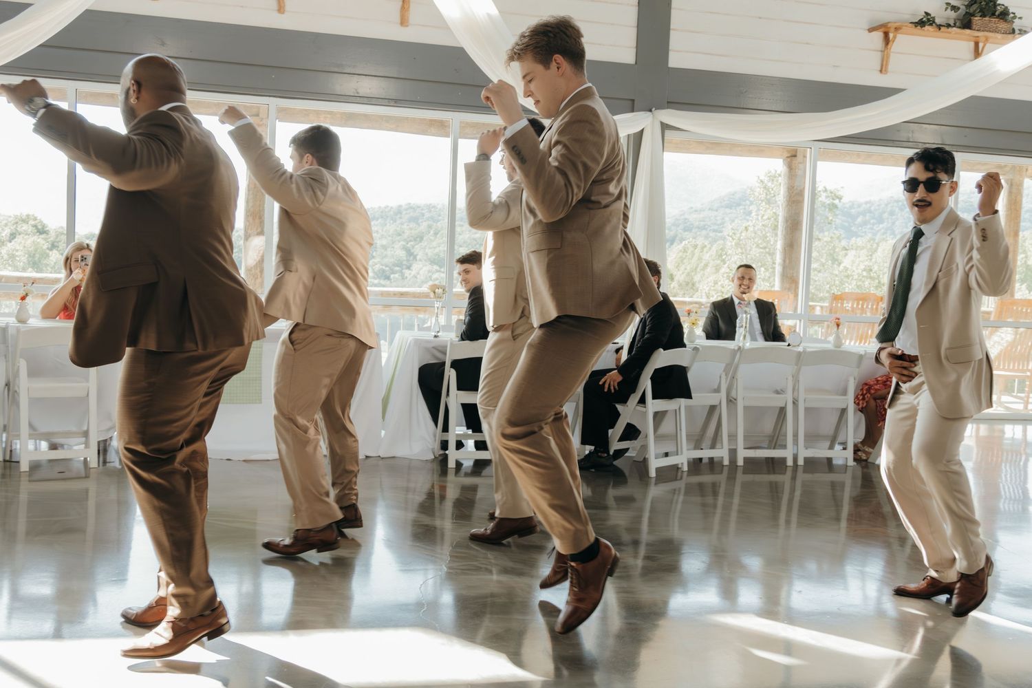 Wedding guests dance energetically in a bright reception venue with large windows.