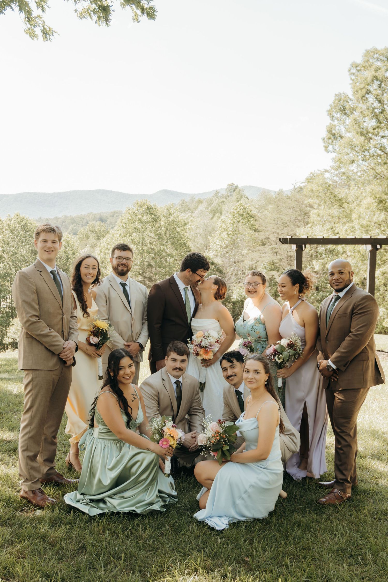 Wedding party in sage green and brown attire poses together on a hilltop with mountain views.