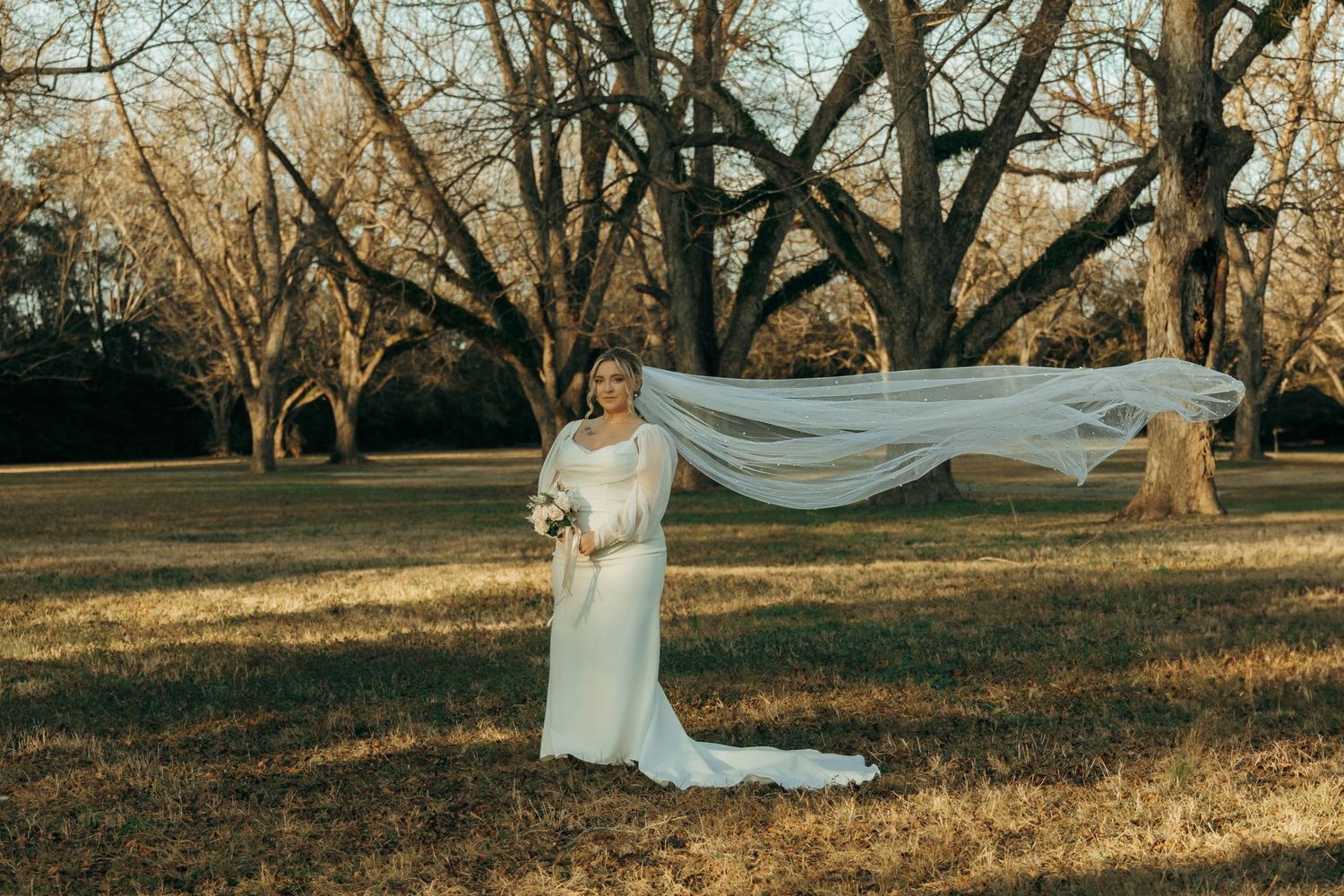 Long wedding veil billows dramatically against backdrop of bare winter trees.