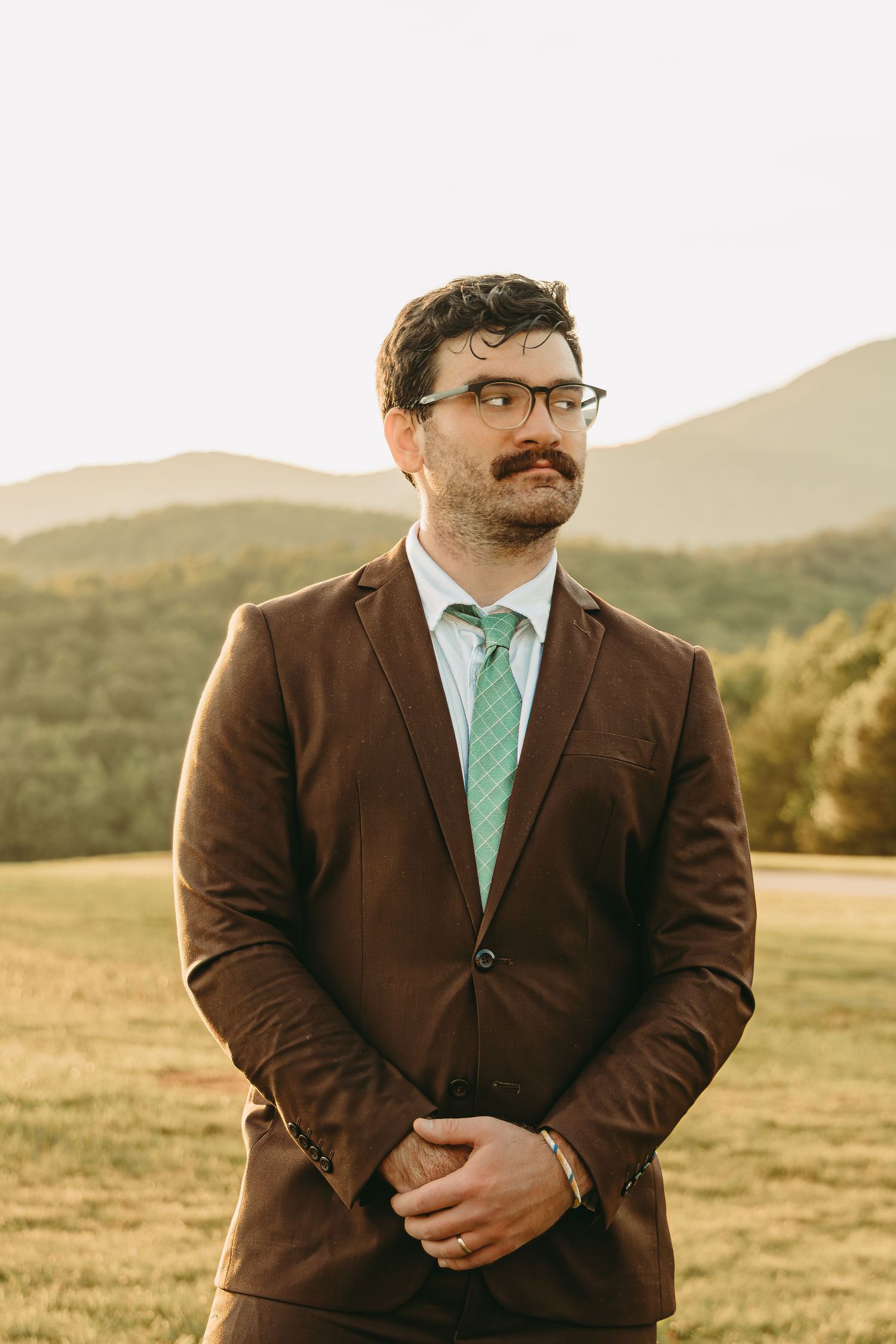 A groom in a brown suit with mint green tie poses in a mountain field at sunset.