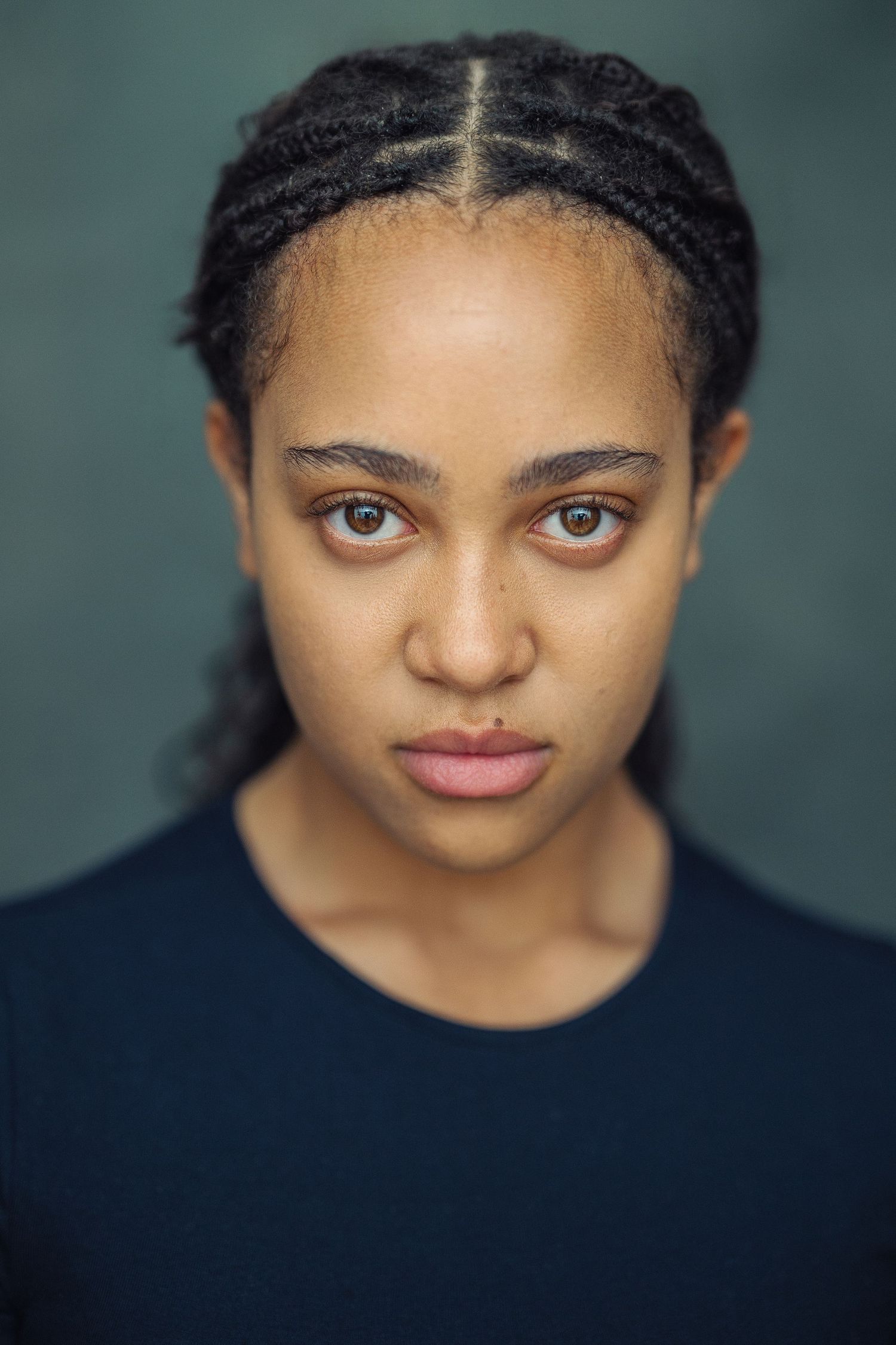 Close-up portrait of a young woman with braided hair and amber eyes wearing a navy blue top against a dark background.