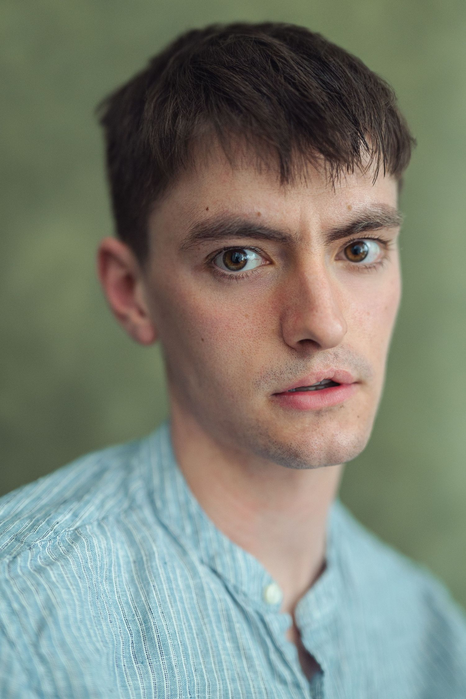 Young man with dark hair and brown eyes wearing a light blue striped shirt, posing against a green background.