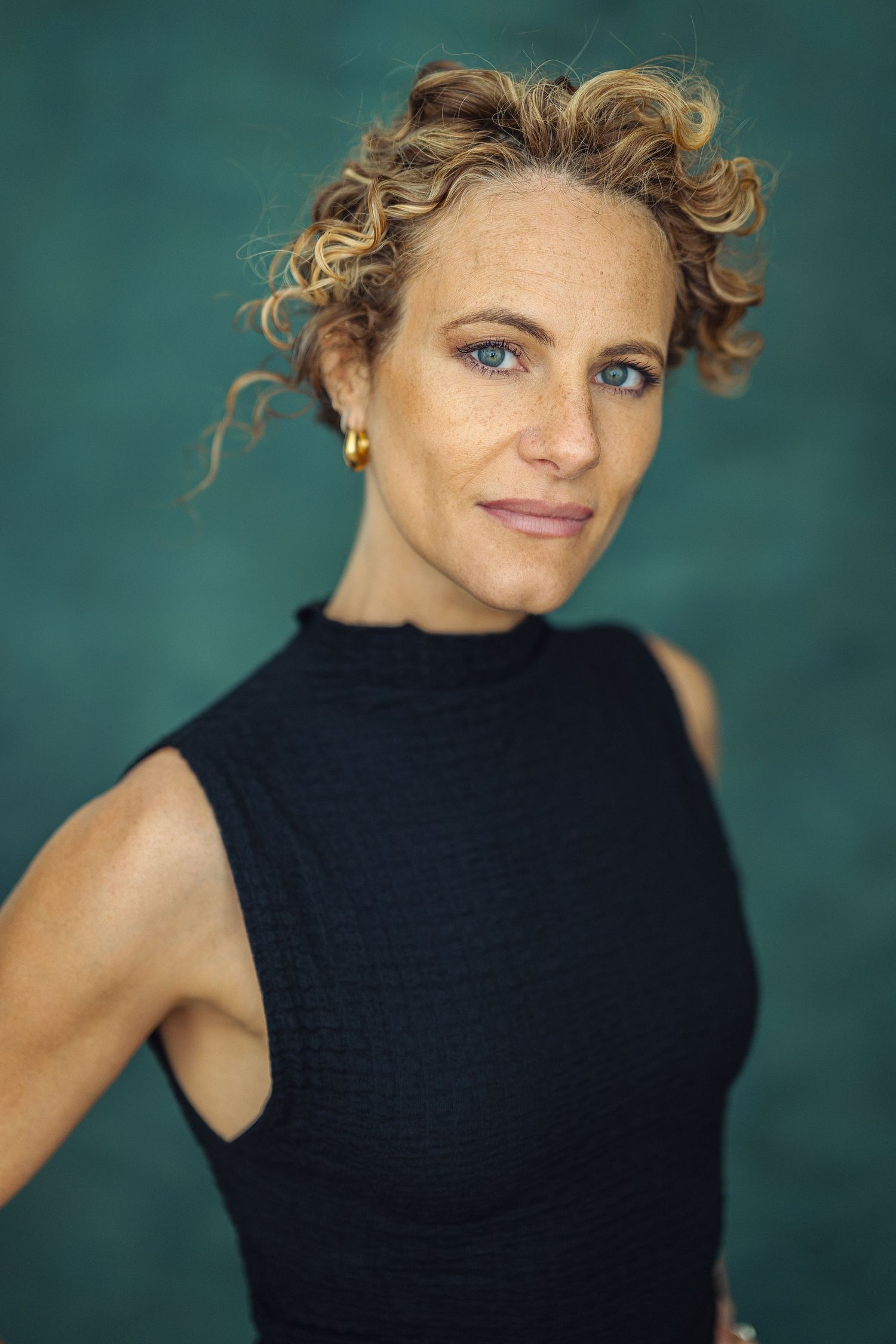 Woman with curly blonde hair and blue eyes wearing a black sleeveless top poses against a teal background.