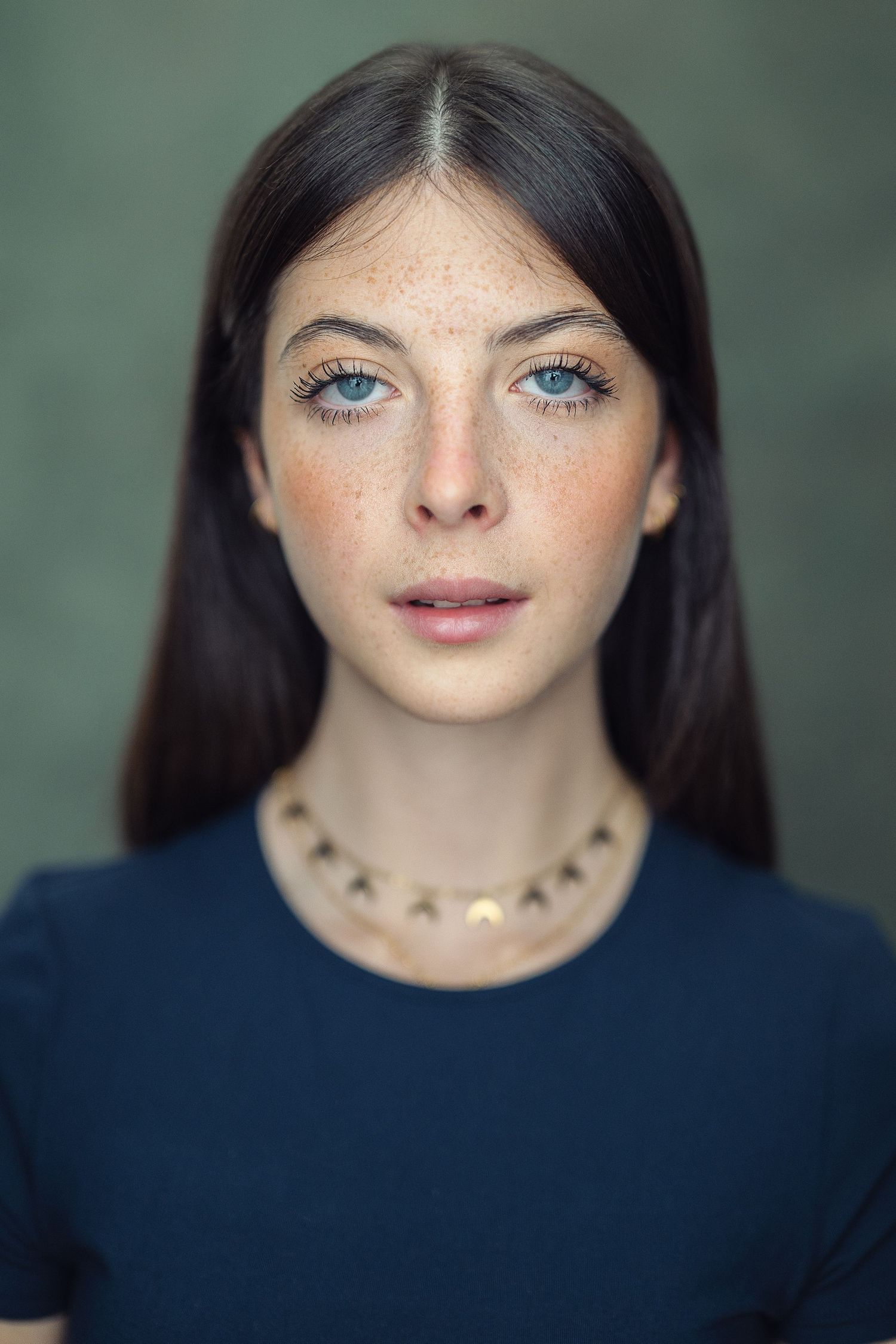 Young woman with blue eyes, freckles, dark hair, gold necklace, and navy shirt poses for a portrait.