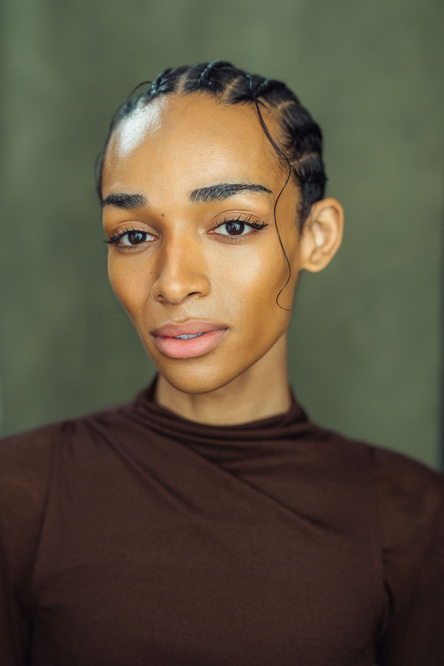 Young Black woman with braided hair and subtle makeup wearing a brown turtleneck top against an olive background.