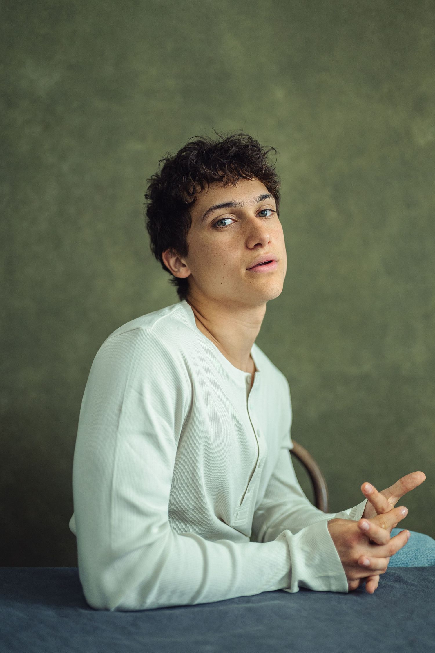 Young person with curly hair in white shirt sits at table, looking up thoughtfully against green backdrop.