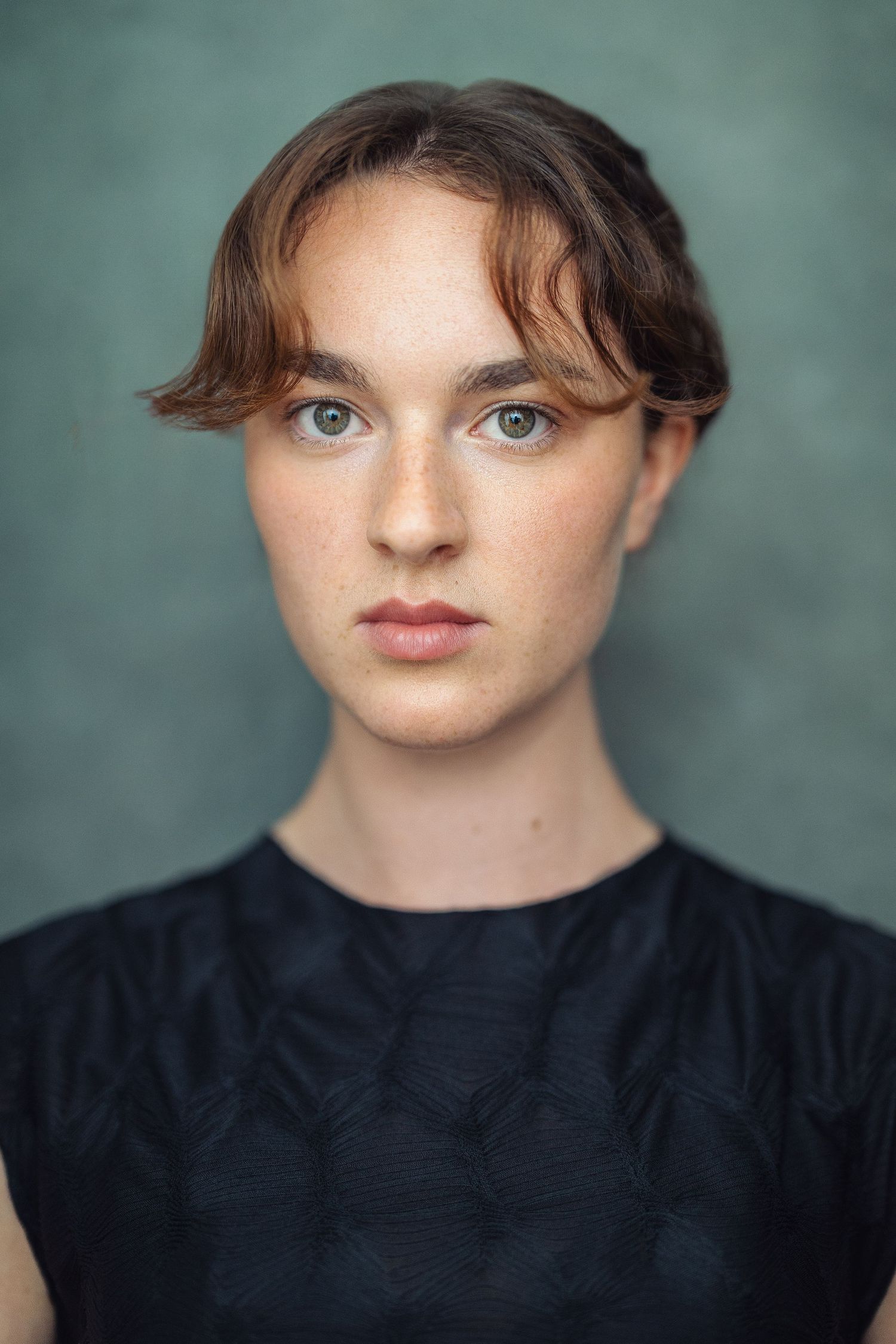 Young woman with short brown hair, green eyes, and freckles wearing a dark patterned top against a teal background.