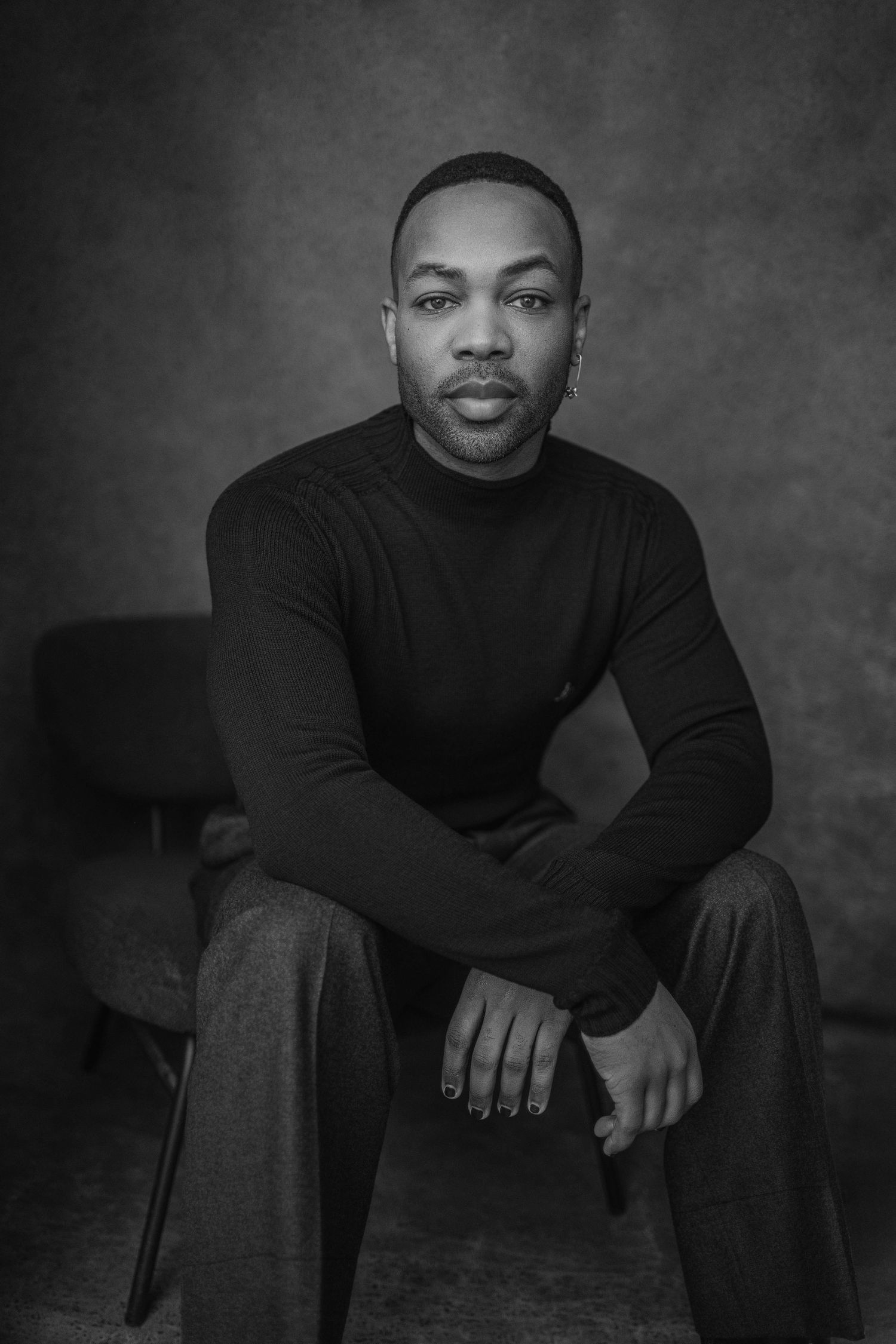 Black and white portrait of a person in a turtleneck sweater sitting on a chair against a dark background.