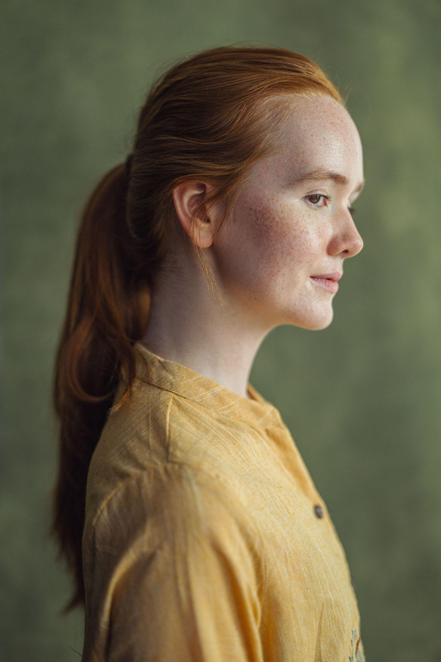 Profile portrait of a young redhead woman with freckles wearing a yellow linen shirt against a green background.