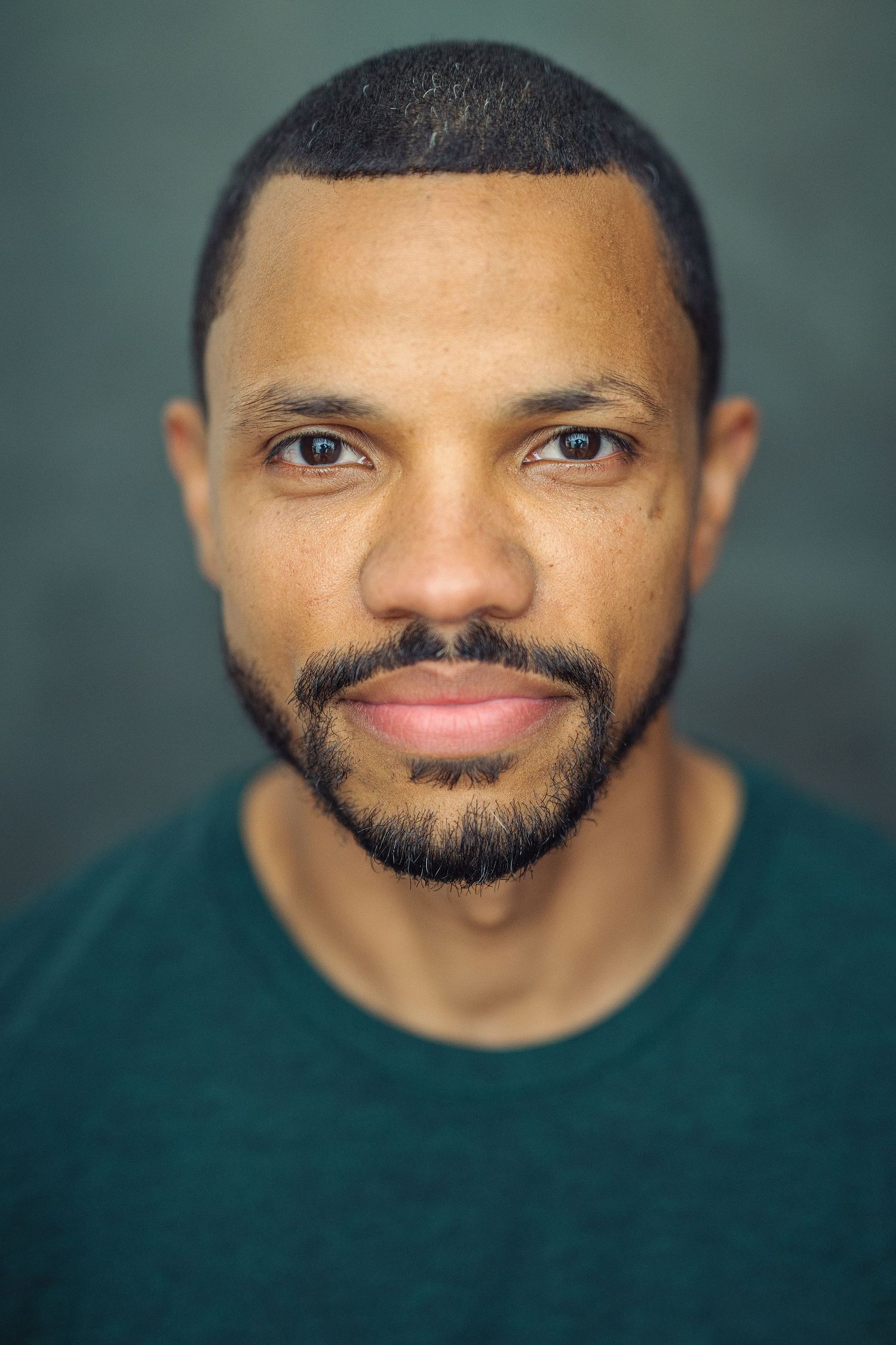 Close-up portrait of a bearded Black man in a teal shirt against a dark green background.