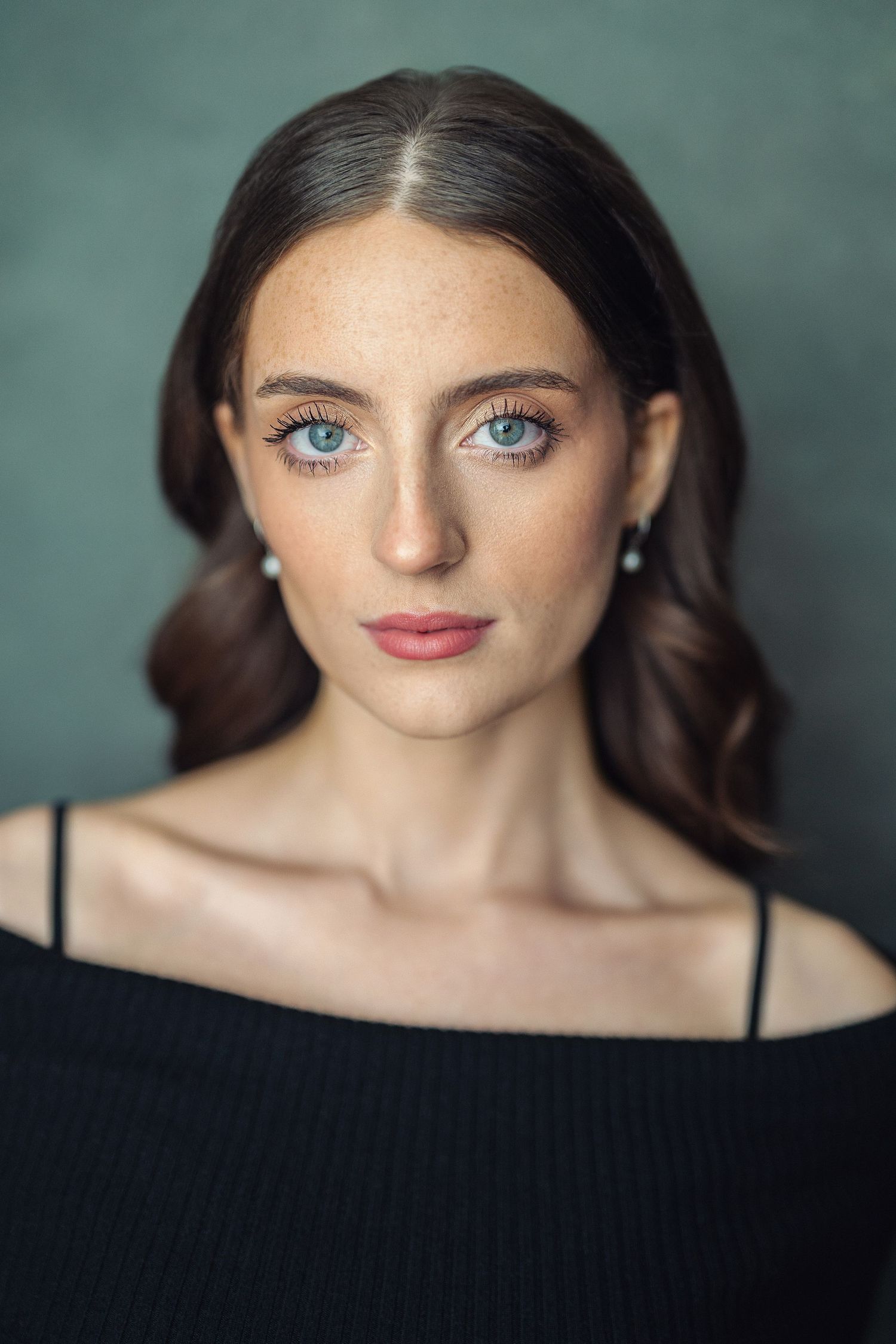 Young woman with blue eyes, freckles, dark wavy hair, wearing black off-shoulder top and pearl earrings against green background.