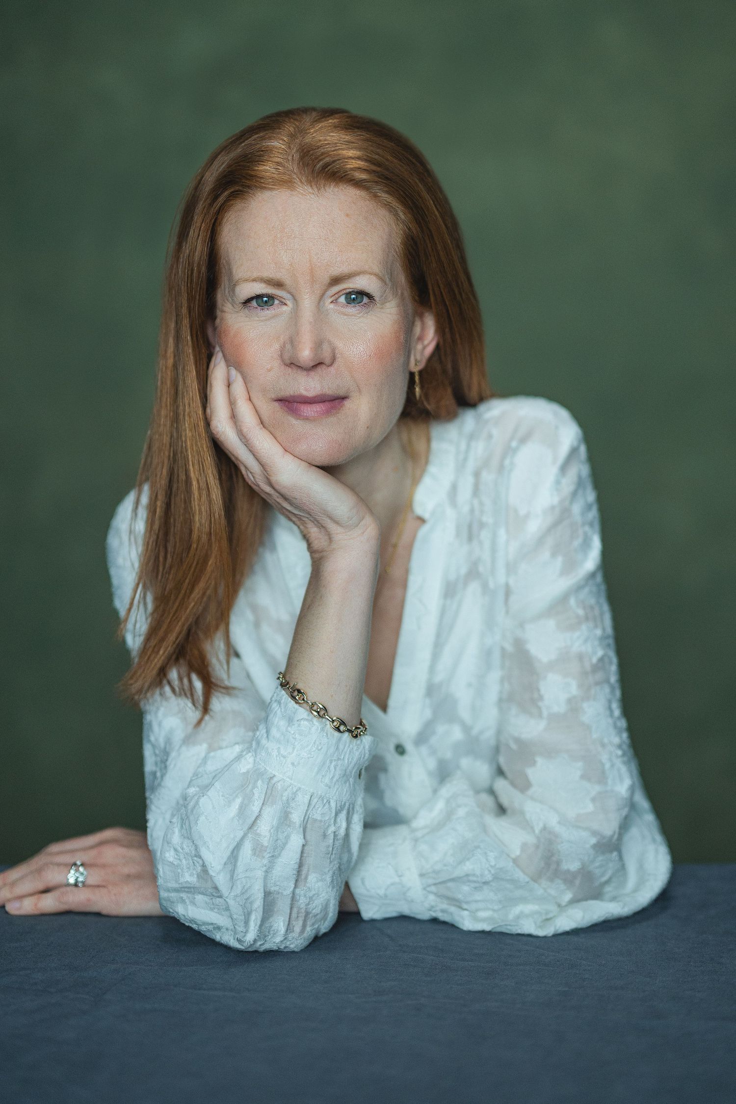 A red-haired woman in a white lace blouse rests her chin on her hand, posing thoughtfully against a green background.