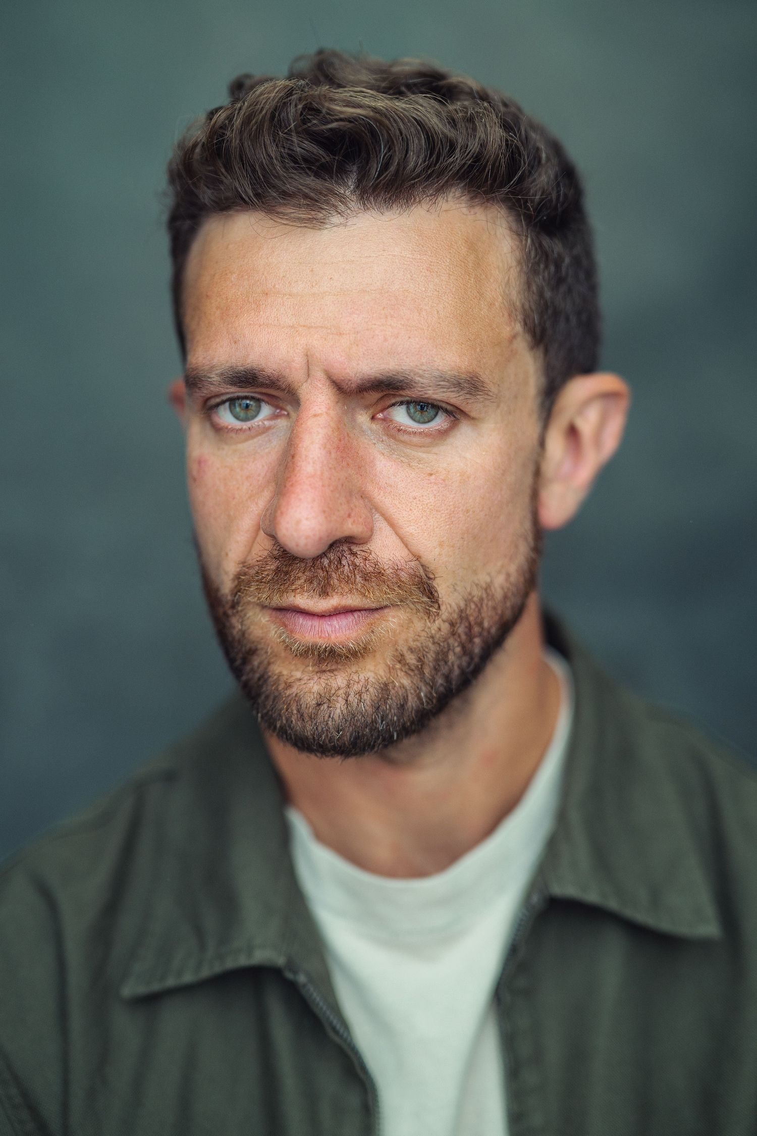 Close-up portrait of a bearded man with blue eyes wearing a green shirt against a dark background.