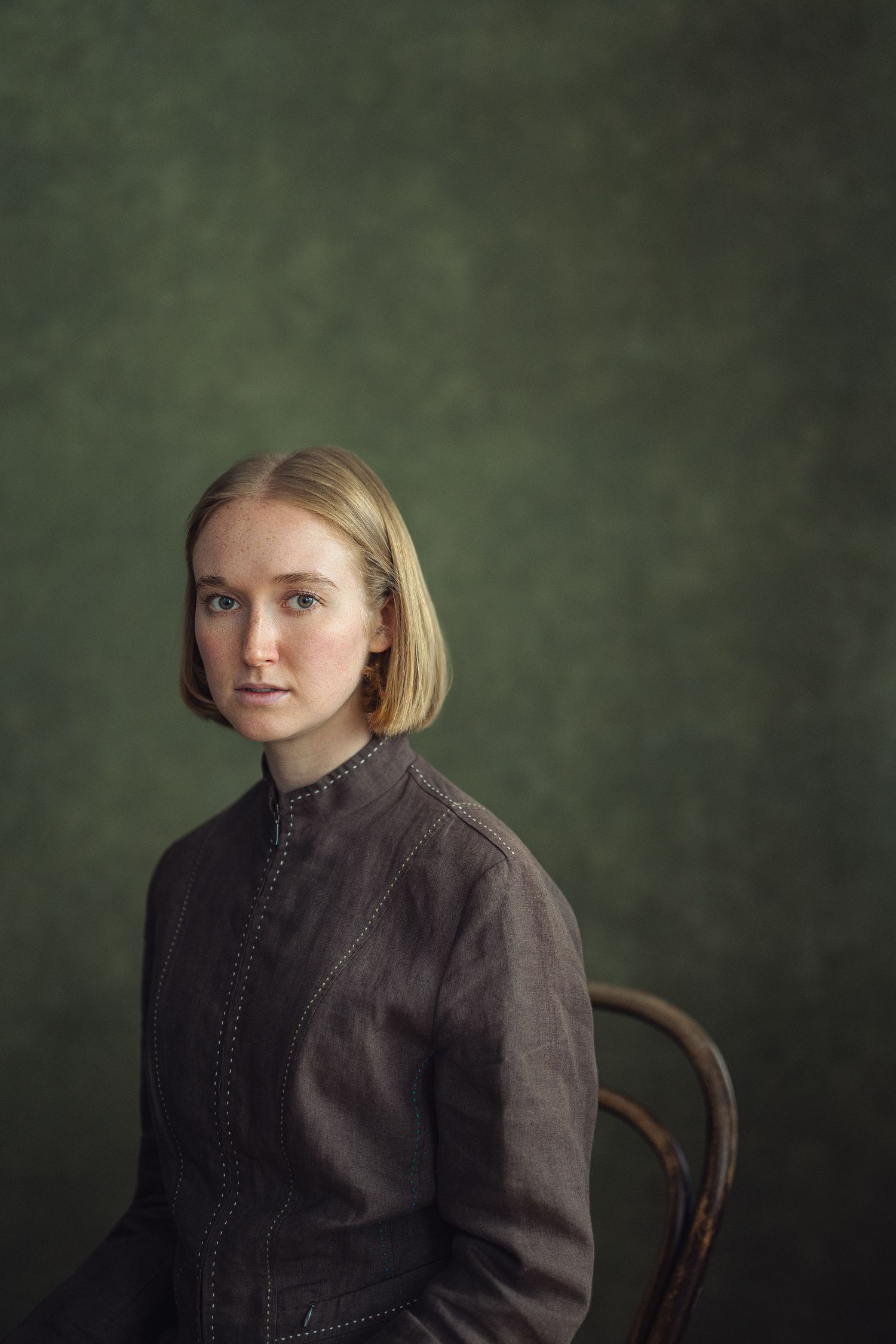 Young blonde woman in gray jacket seated on wooden chair against dark green studio backdrop.