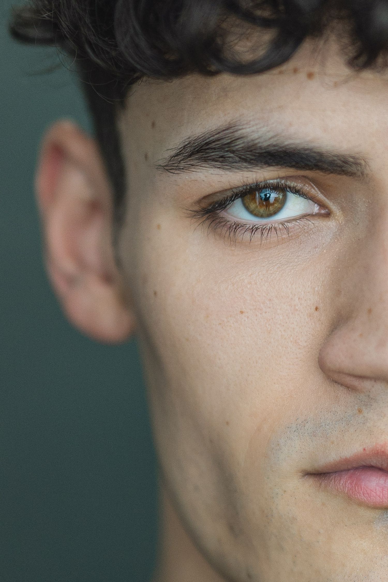 Extreme closeup portrait showing half a face with striking hazel eyes and dark curly hair against a teal background.