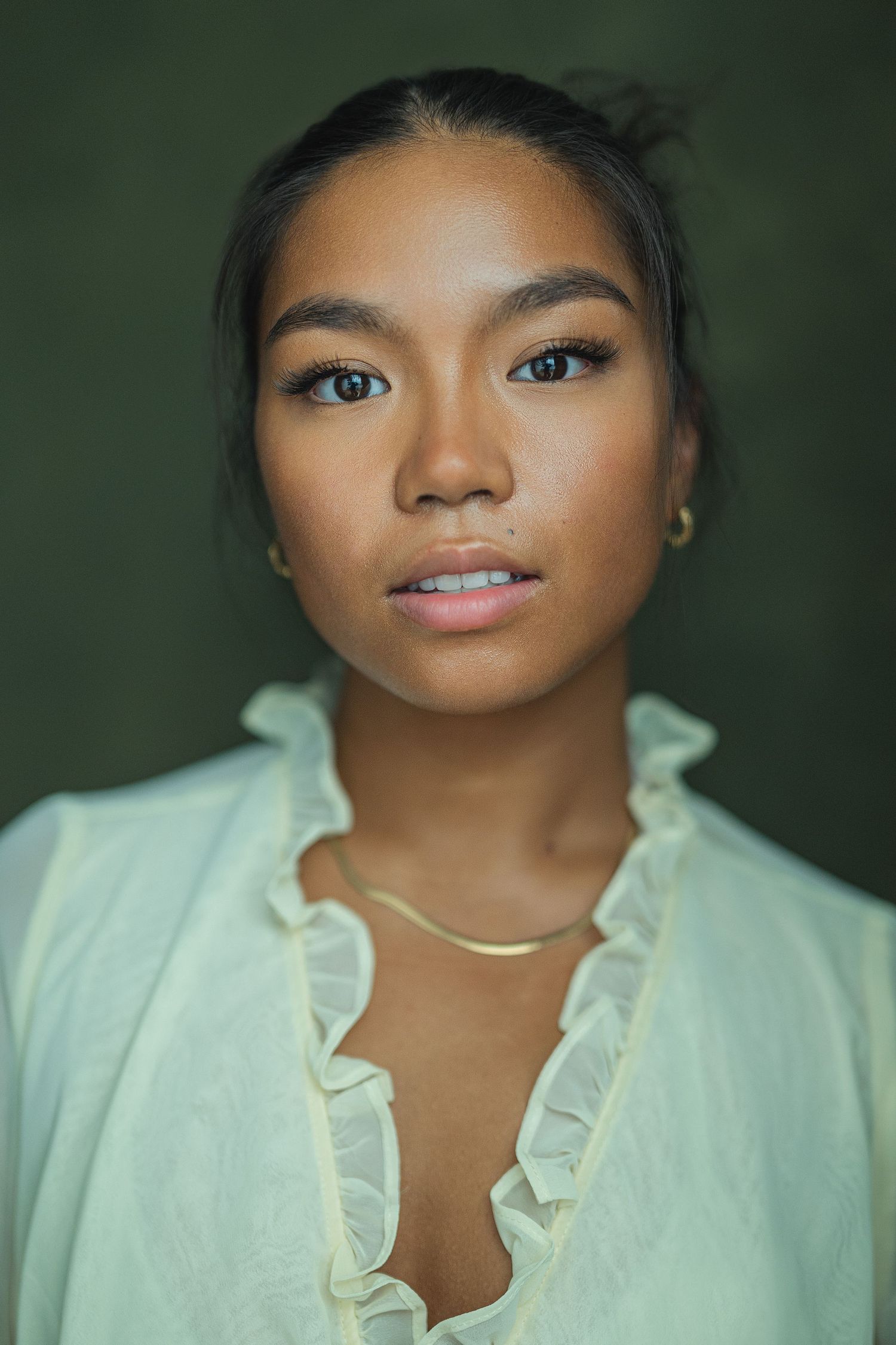 Portrait in white ruffled blouse against dark green background with soft lighting and gold necklace.