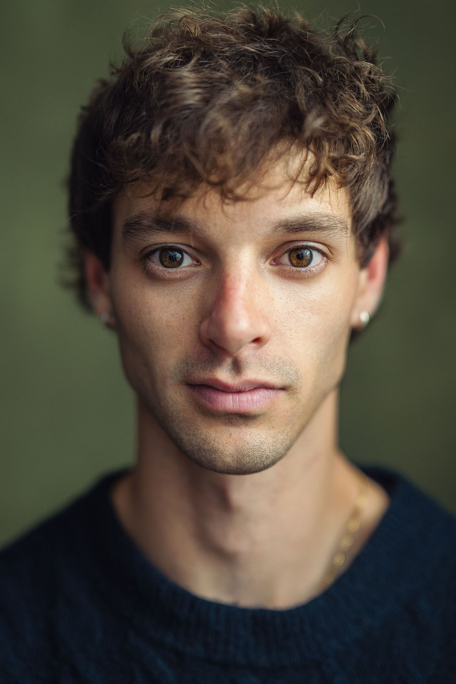 Young man with curly brown hair, brown eyes, small earring, and light stubble wearing a dark sweater against green background.