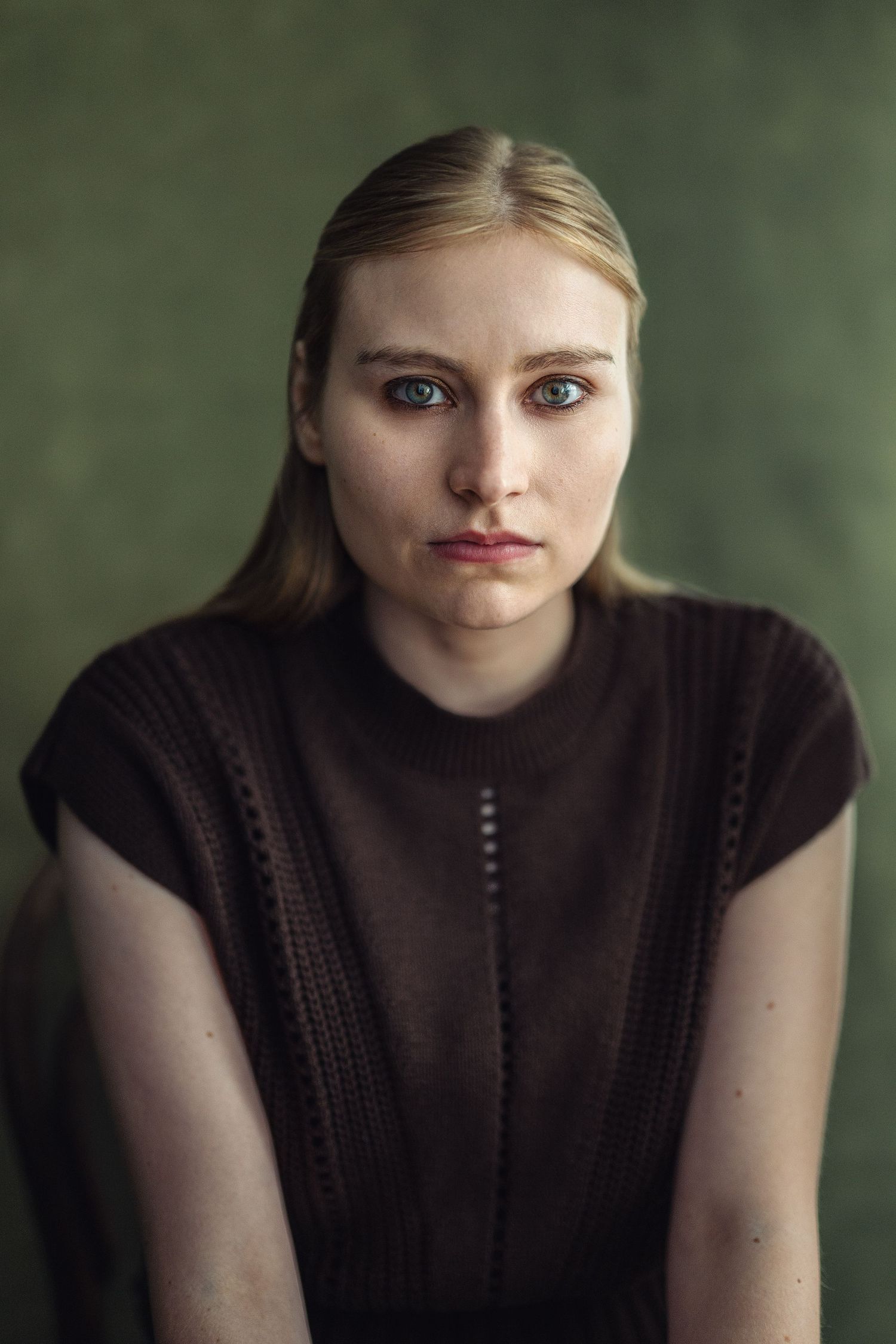 Young blonde woman with green eyes wearing a dark brown knit sleeveless top, posed against a muted green background.