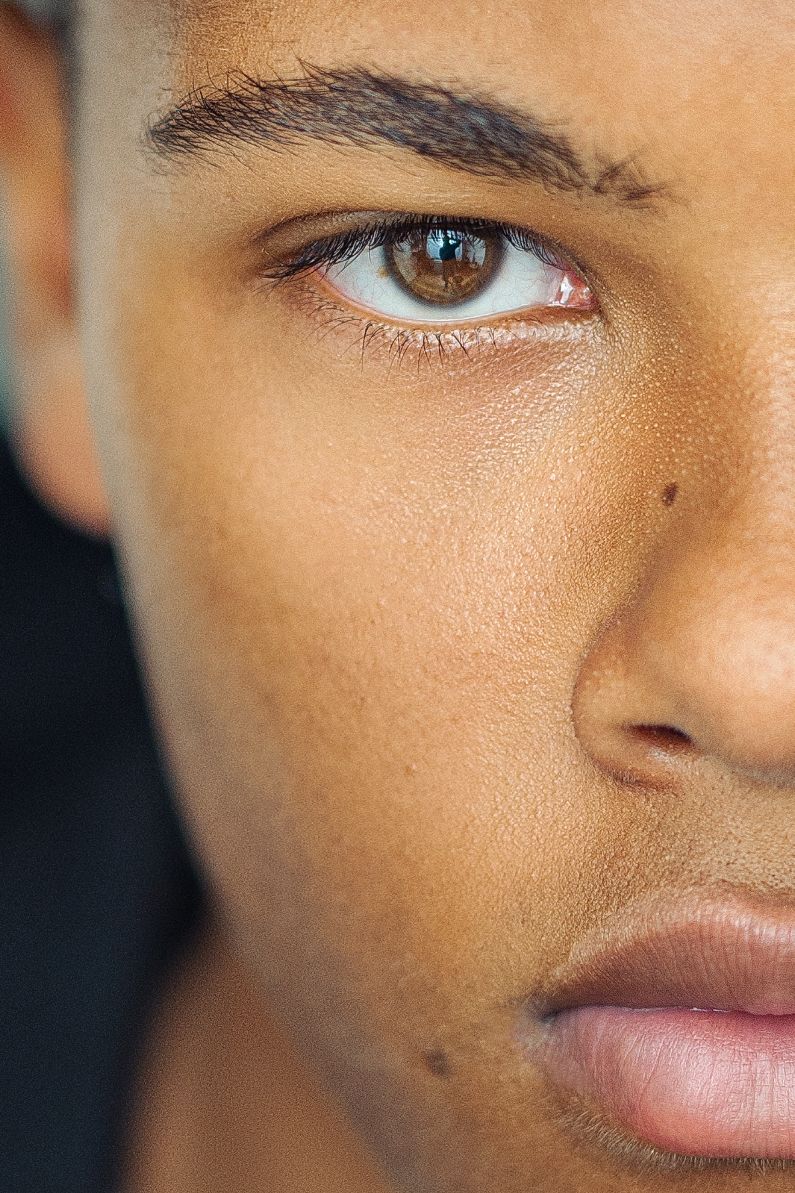 Close-up portrait of a young person with brown eyes, smooth skin, and defined eyebrows against a dark background.