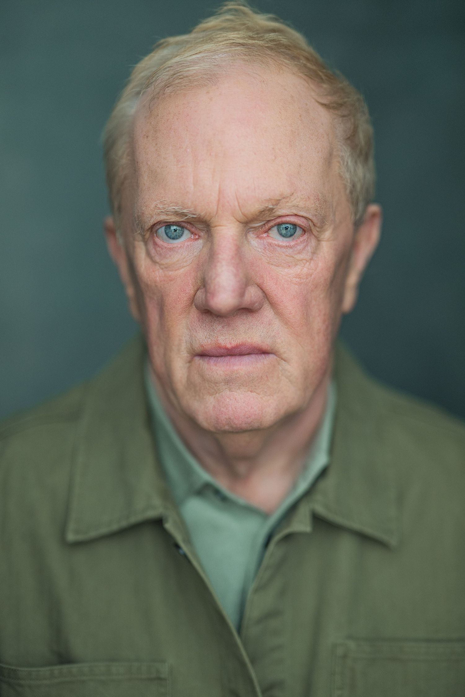 A close-up portrait of an older man with blue eyes wearing an olive green jacket against a teal background.