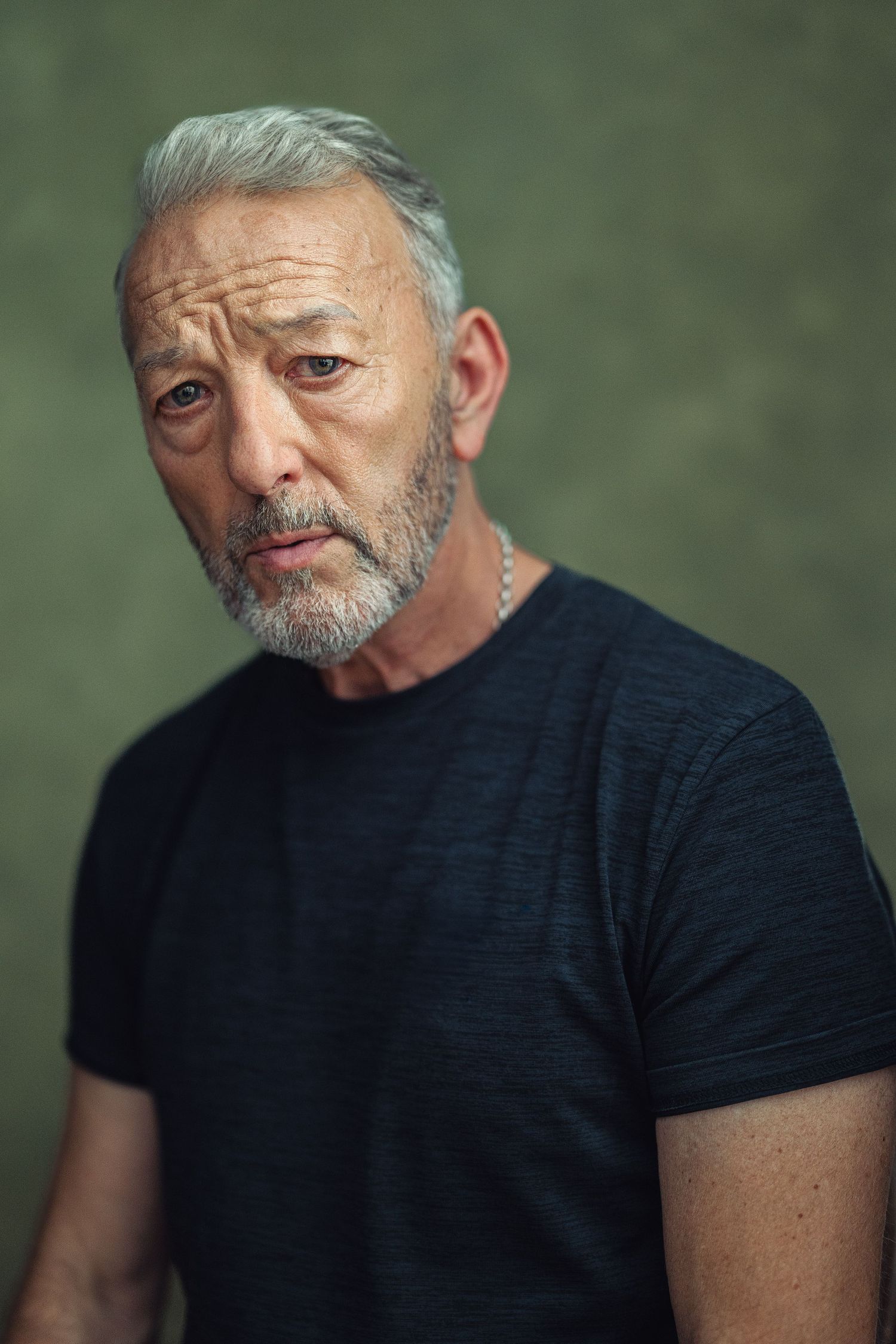 Older man with gray hair and beard wearing a black t-shirt, posing against a dark green background.