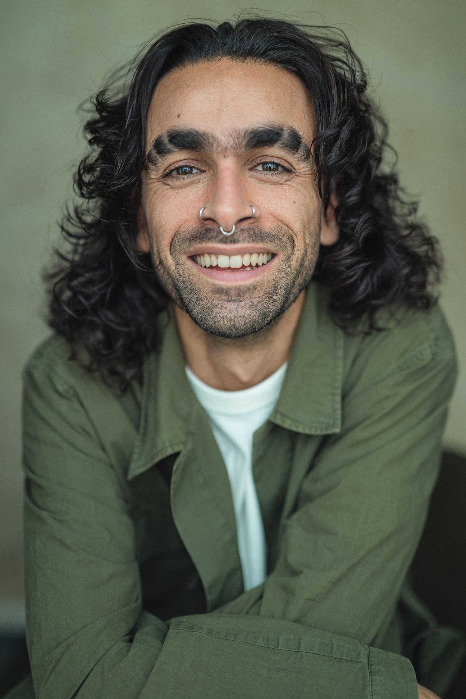 Portrait of someone with long dark wavy hair, nose piercing and bright smile wearing a green jacket and white t-shirt.
