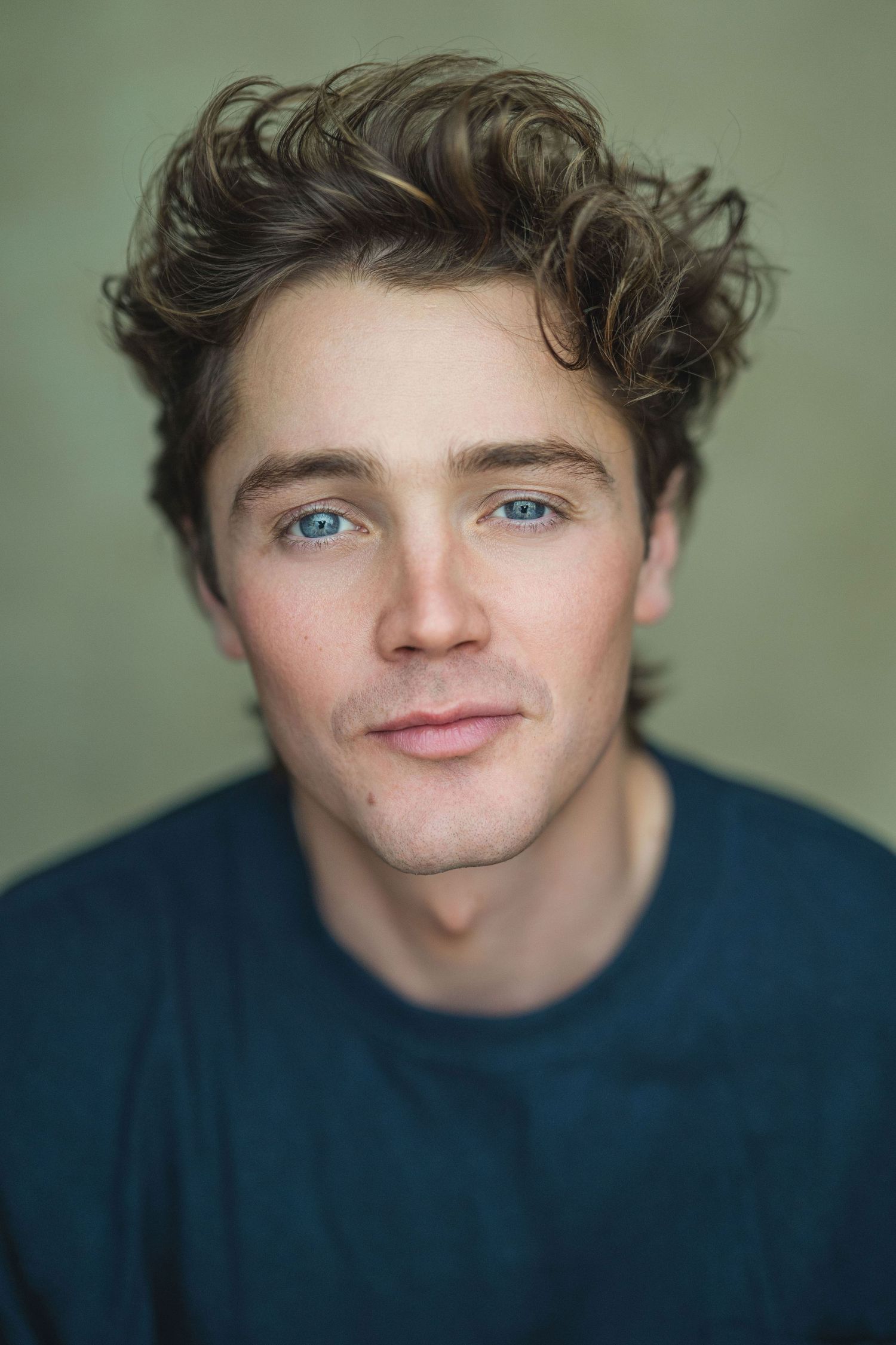 Portrait of a young person with wavy brown hair and blue eyes wearing a navy sweater against a green background.