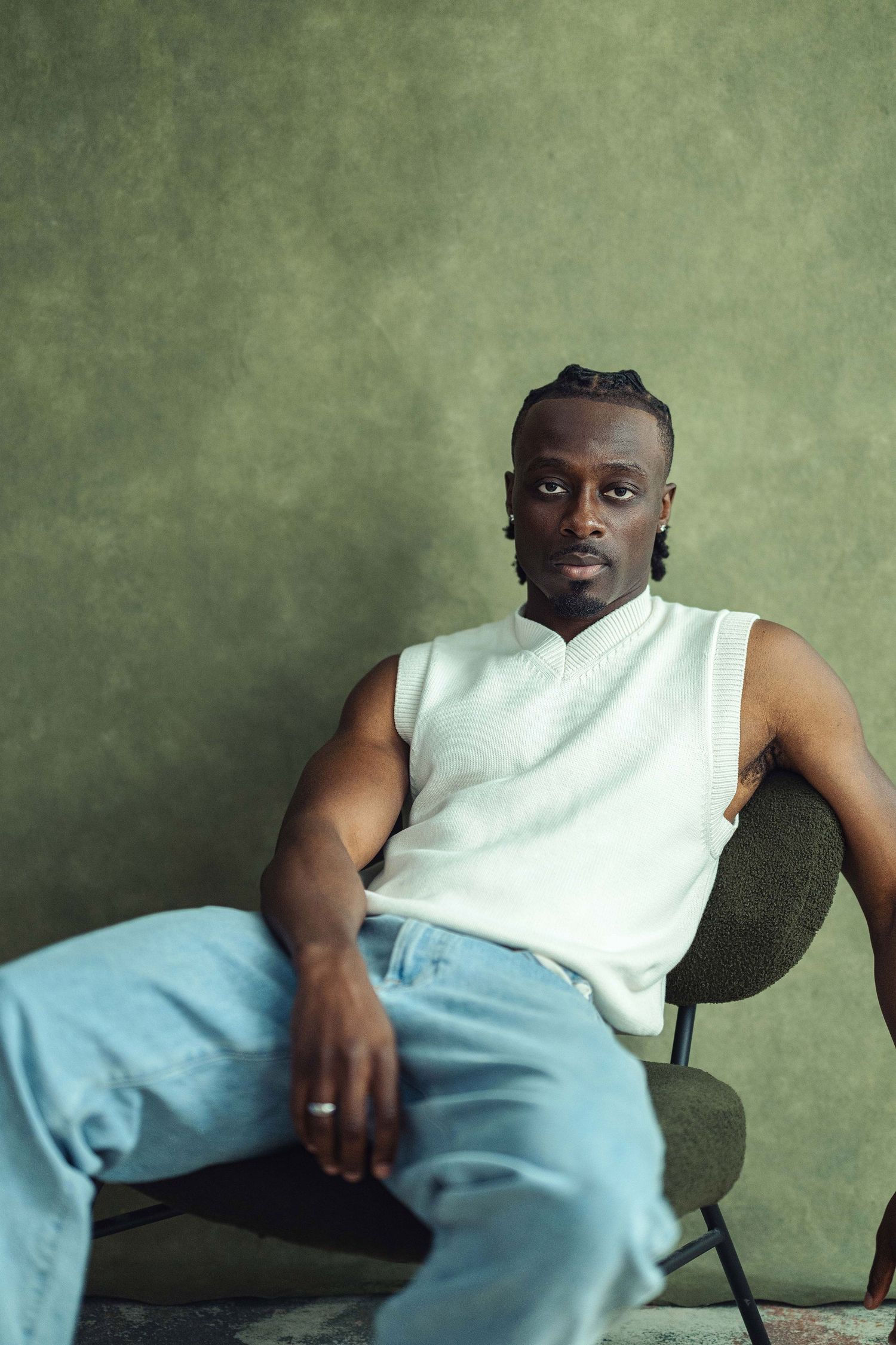 A young Black man in a white sleeveless top and light blue jeans sits relaxed in a chair against an olive green wall.