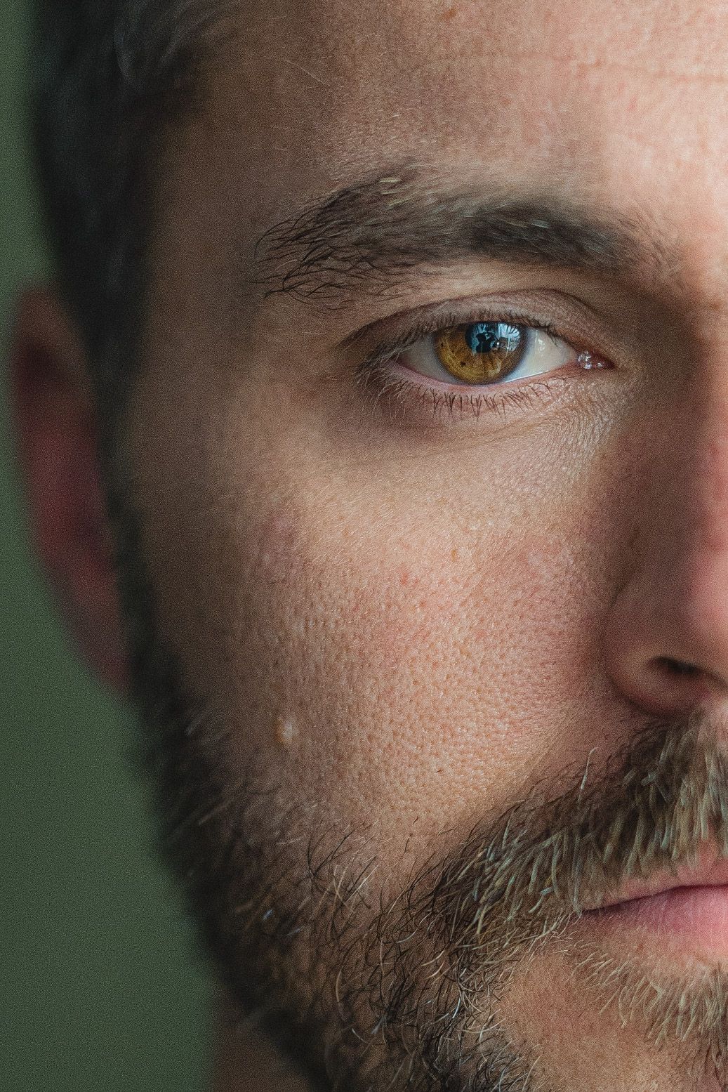 Extreme close up macro photograph showing a striking brown eye and partial bearded face against a green background.