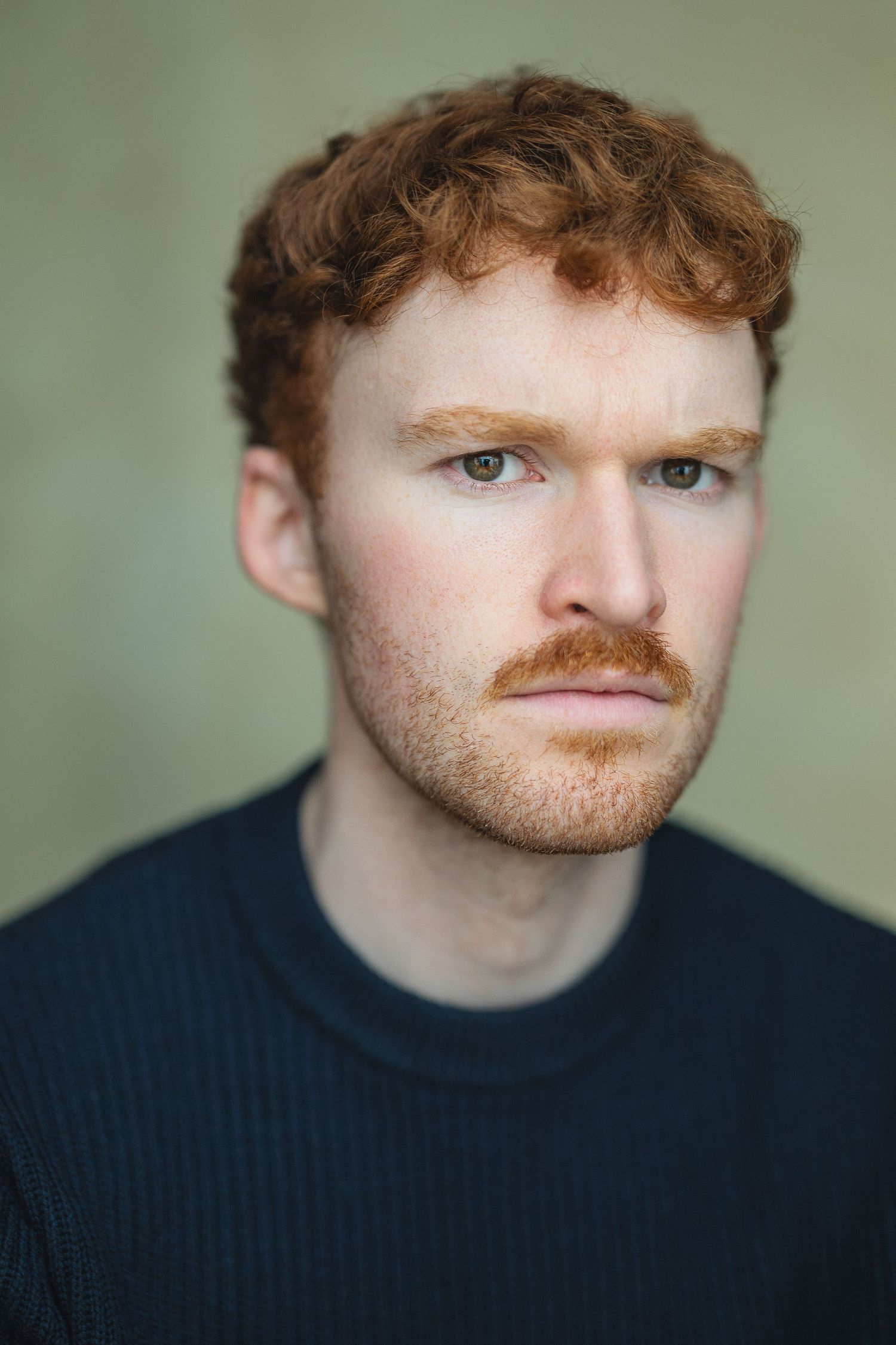 Portrait of someone with curly red hair and a ginger mustache wearing a dark sweater against a neutral background.