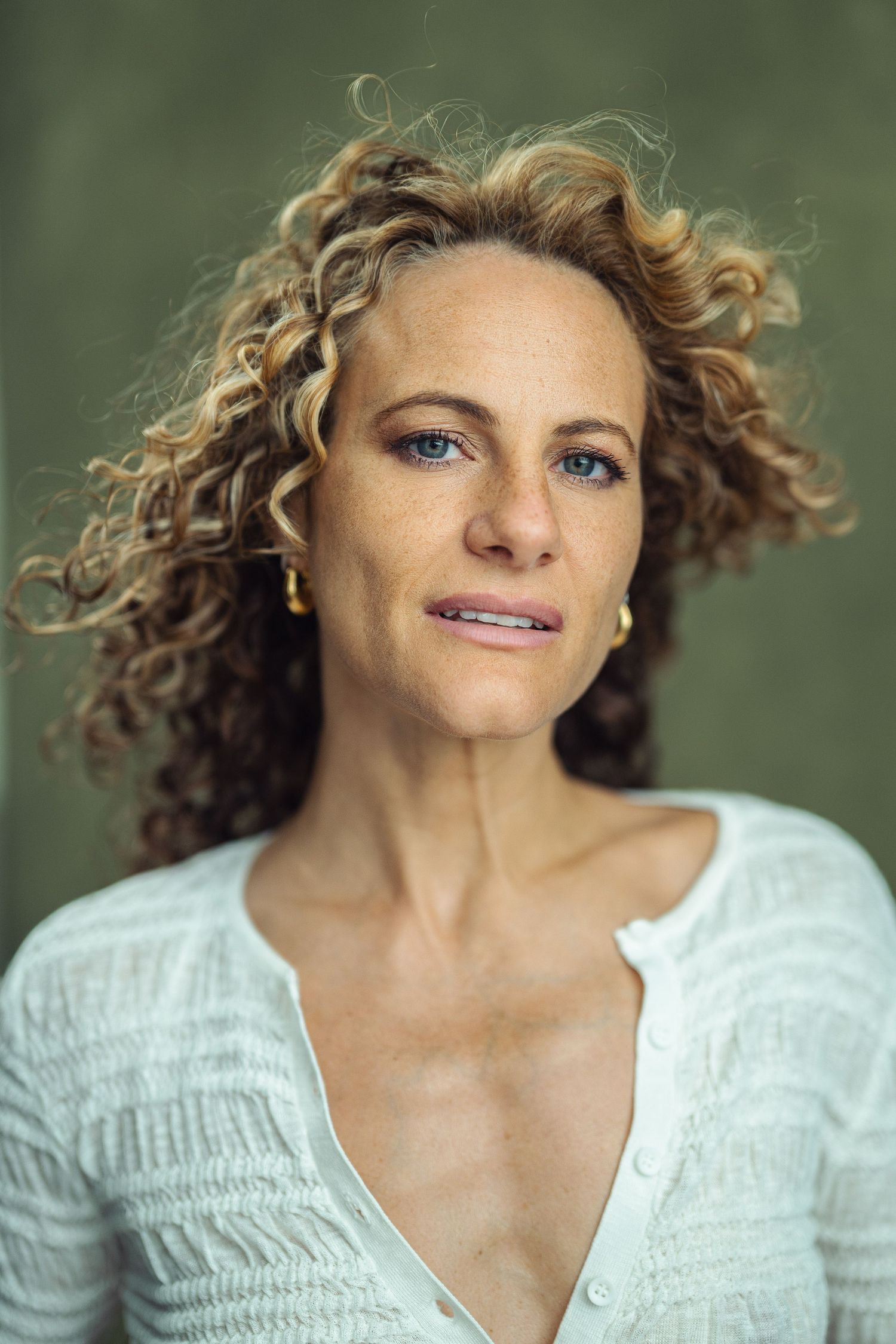 Portrait of a curly-haired blonde woman with blue eyes and freckles wearing a white top and gold earrings.